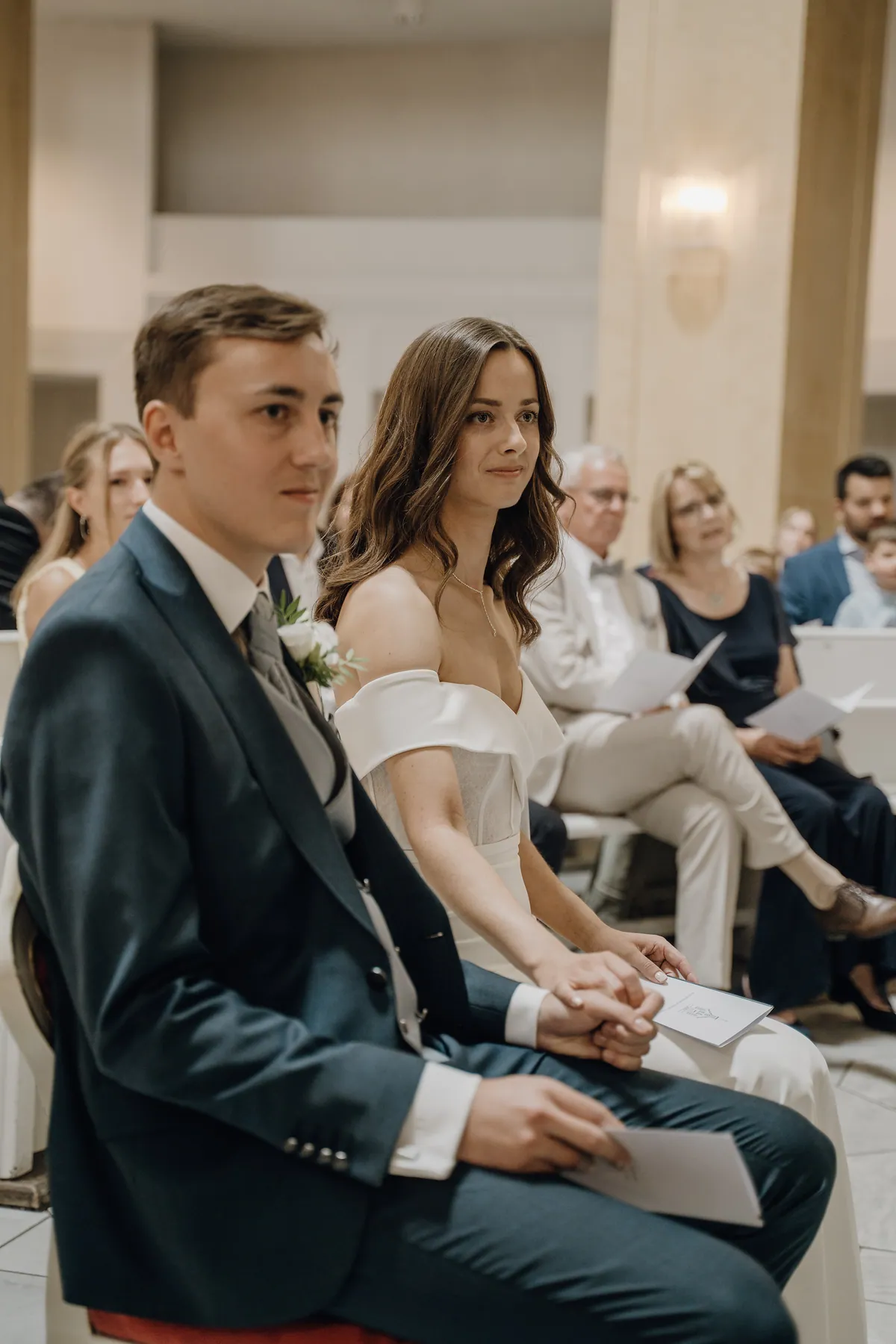 Luisa und Jonas sitzen nebeneinander, ihre Hand liegt auf seiner, beide halten ein Programmheft auf dem Schoß, Luisa blickt mit leichtem Lächeln nach vorn, hinter ihnen unscharf die Gäste.