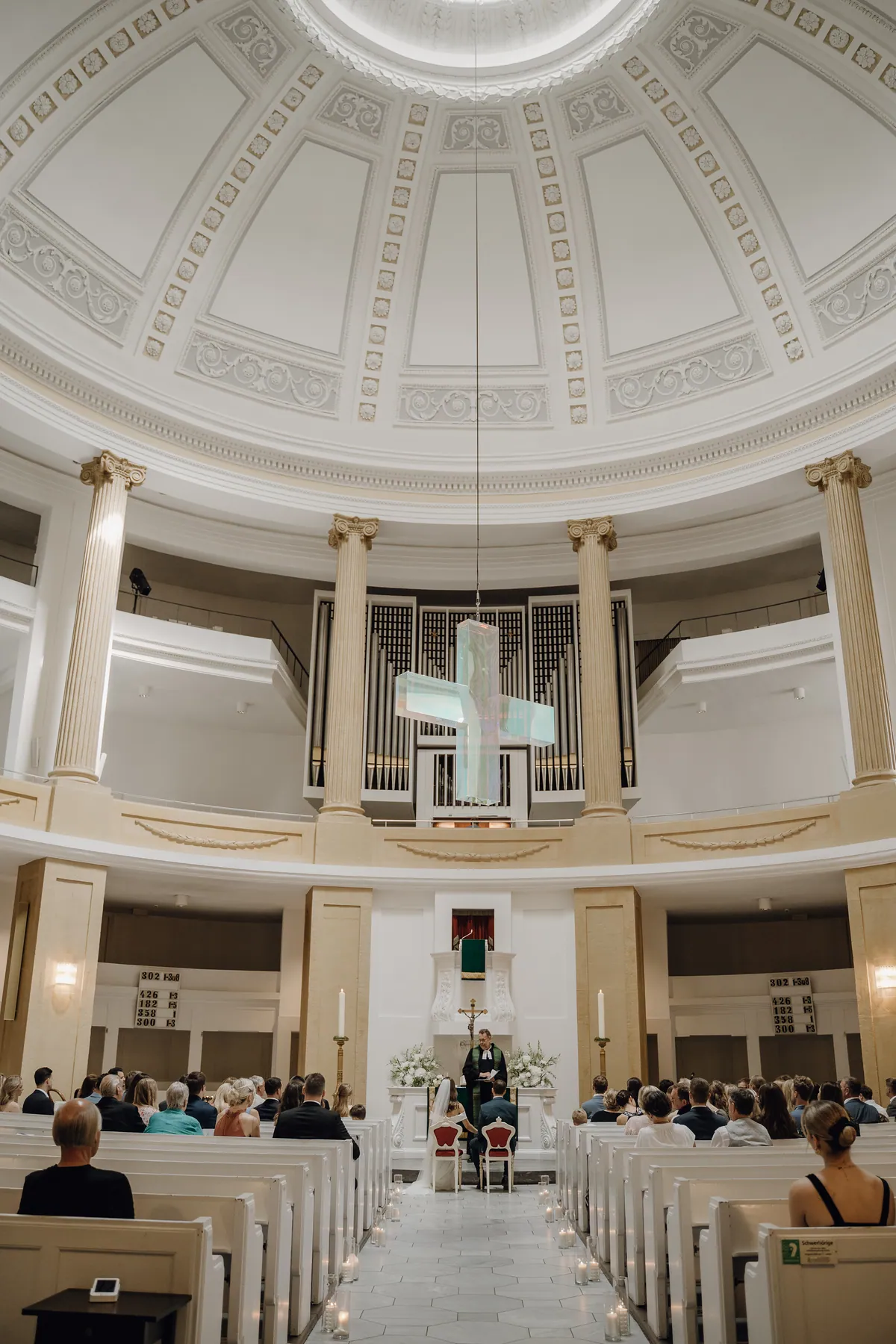 Unter der weißen Stuckkuppel sitzen Luisa und Jonas, von hinten gesehen, auf zwei roten Stühlen vor dem Altar, ein Pfarrer im schwarzen Talar steht vor ihnen, die Gäste füllen die Bänke beiderseits des Gangs.