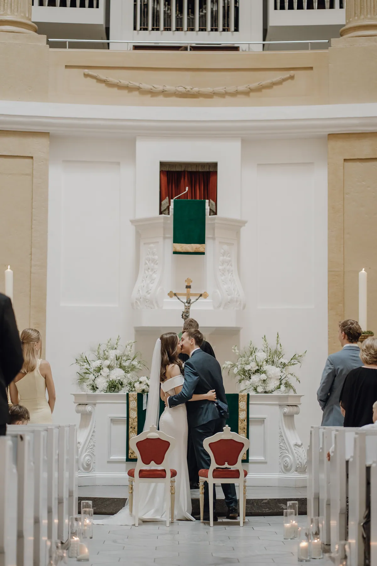 Vor dem Altar mit Kruzifix und zwei üppigen weißen Blumengestecken küssen sich Luisa und Jonas hinter den beiden roten Stühlen, darüber die Kanzel mit grünem Behang, links und rechts stehen Gäste.