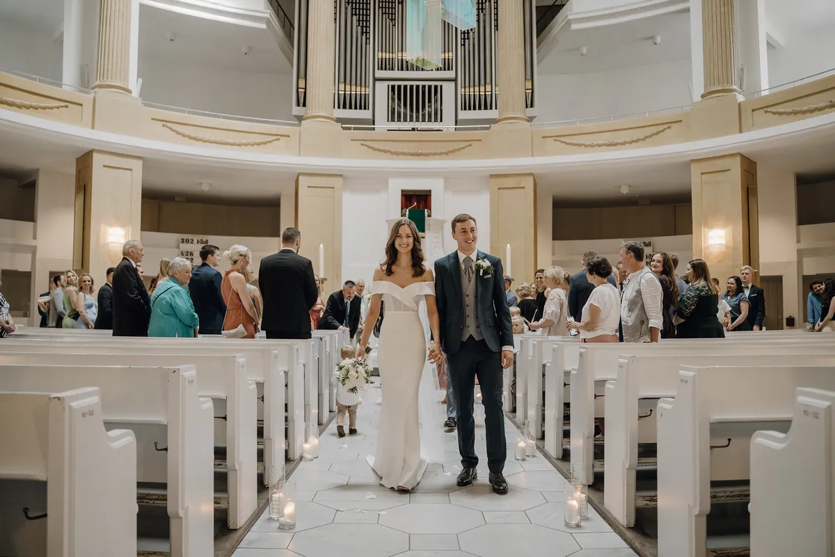 Hand in Hand gehen Luisa und Jonas lächelnd durch den kerzengesäumten Mittelgang auf die Kamera zu, hinter ihnen läuft ein Kleinkind, Gäste stehen in den Bänken, oben die Orgel.