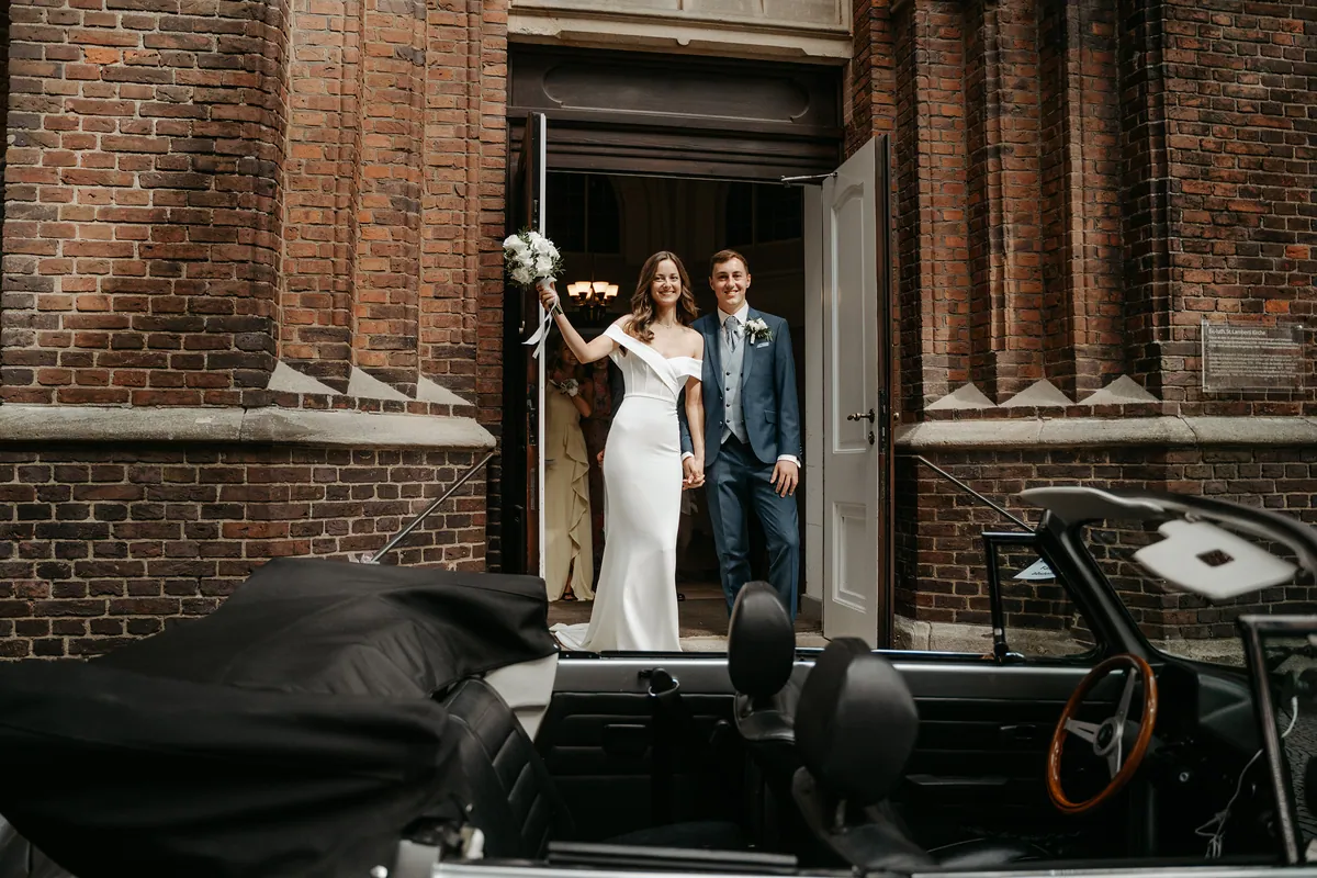 Hand in Hand stehen Luisa und Jonas lächelnd im geöffneten Portal der Backsteinkirche, sie hebt den weißen Brautstrauß in die Höhe, im Vordergrund das offene Cabrio mit Holzlenkrad.