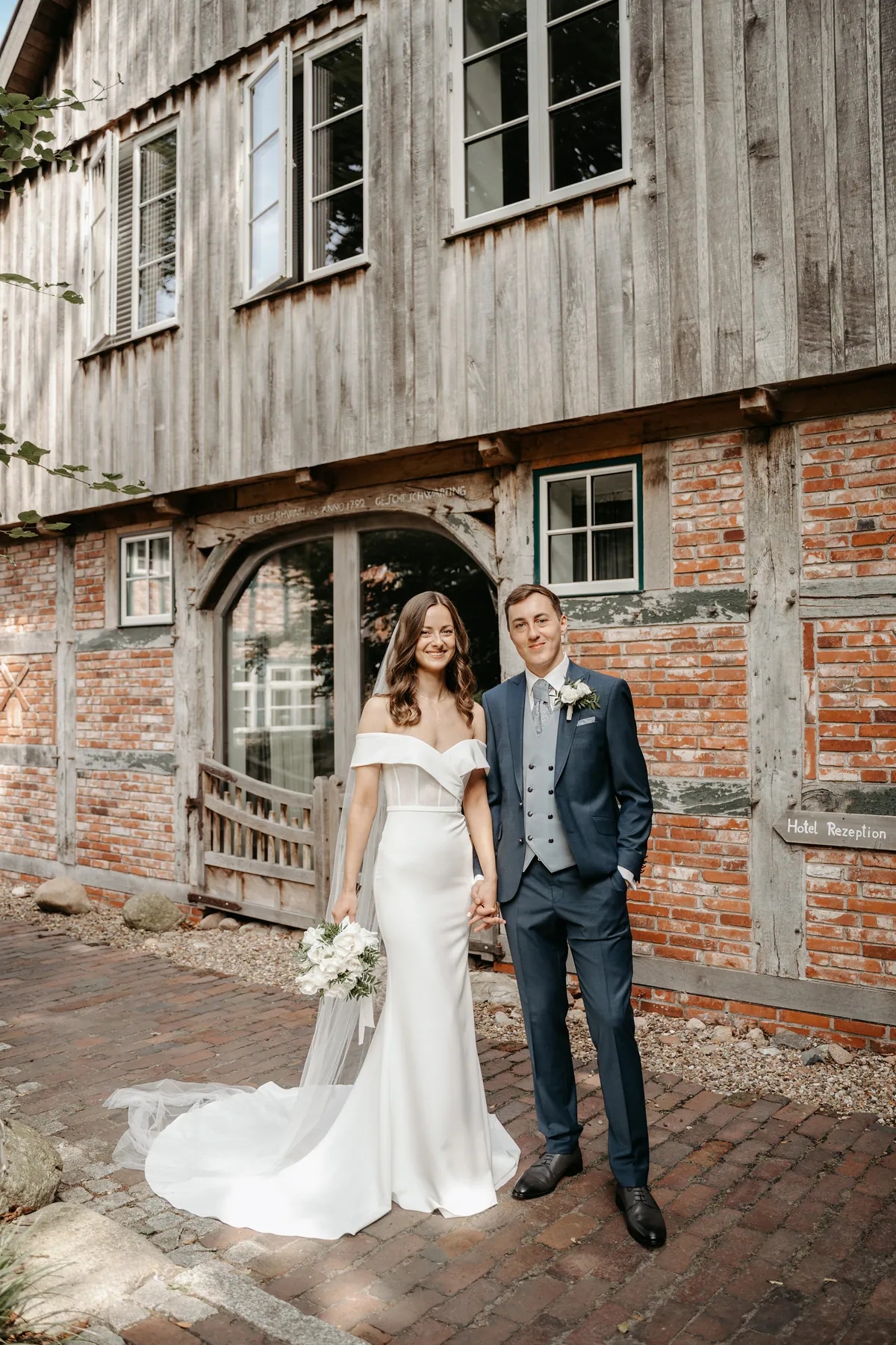 Luisa und Jonas stehen Hand in Hand vor dem Fachwerkhaus und blicken lächelnd in die Kamera, ihr Schleier und die lange Schleppe liegen auf dem Klinkerpflaster, er hat eine Hand in der Hosentasche.