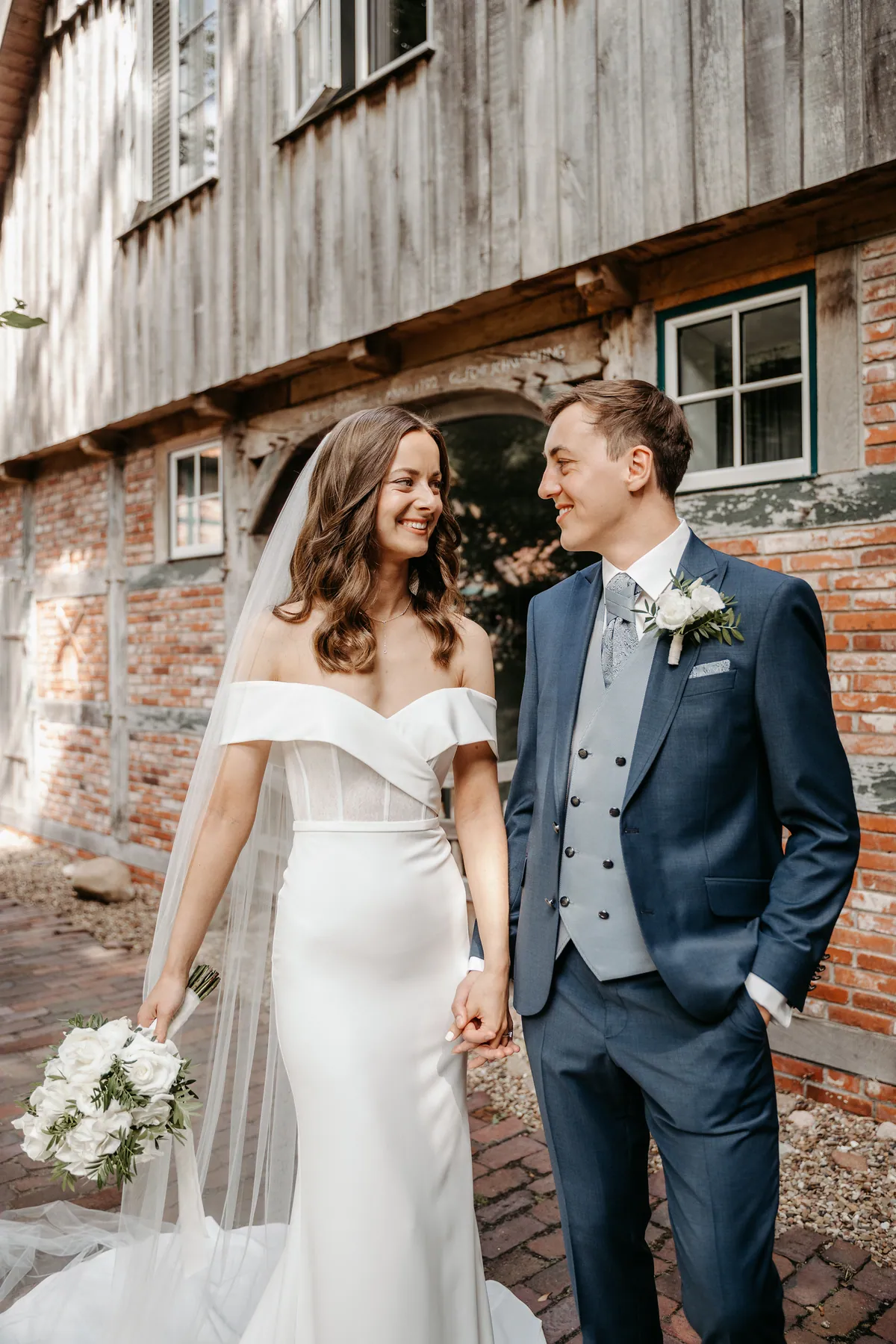 Hand in Hand sehen sich Luisa und Jonas lächelnd an, sie hält den Rosenstrauß locker an der Seite, er trägt eine zweireihige graublaue Weste unter dem dunkelblauen Sakko.