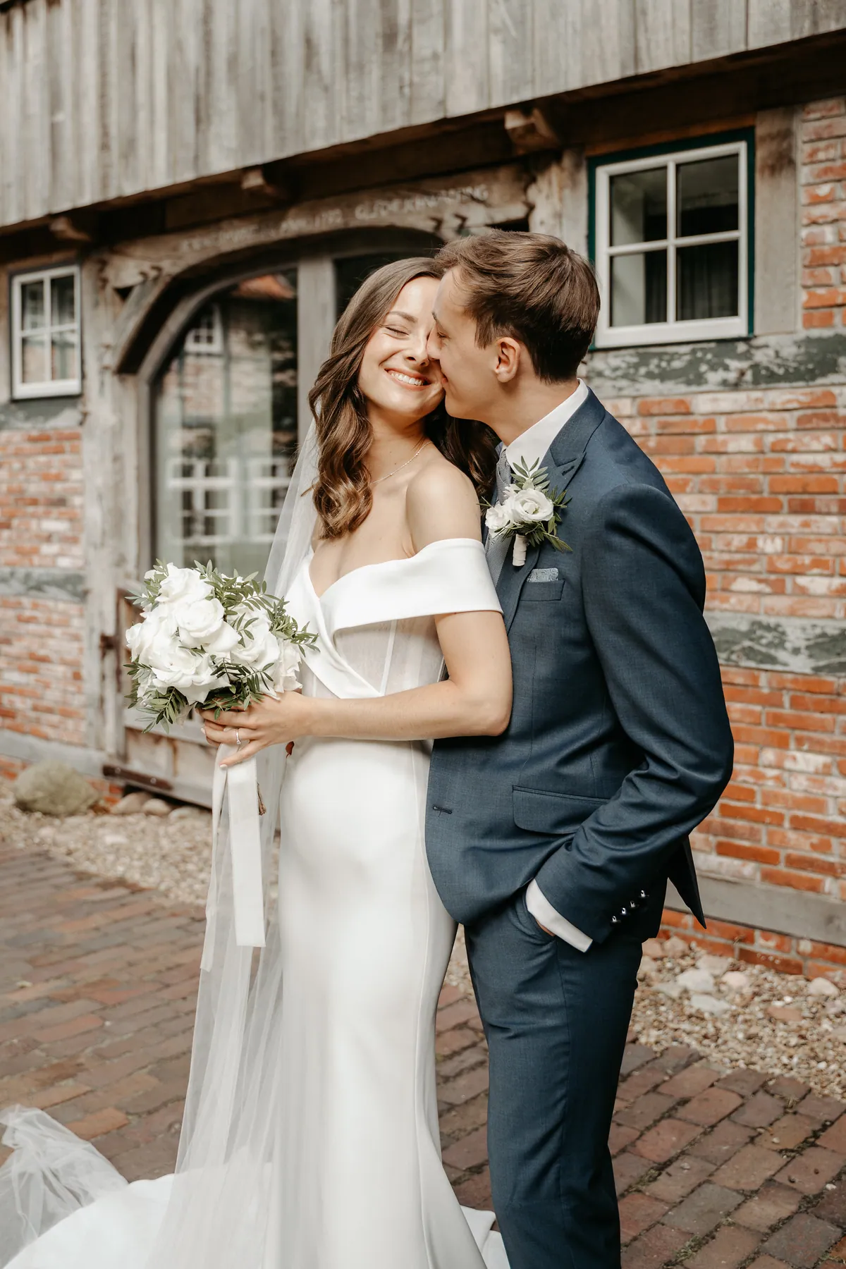 Jonas küsst Luisa auf die Wange, sie lacht mit geschlossenen Augen und hält den Strauß aus weißen Rosen und schmalblättrigem Grün vor sich, dahinter Fachwerk und Backstein.