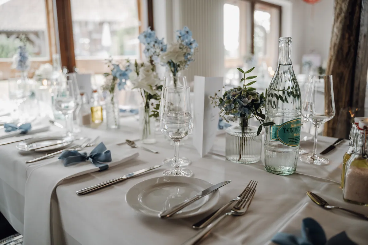 Nahaufnahme eines Gedecks: Teller mit Messer und Gabel, Serviette mit taubenblauer Schleife, Weingläser, ein Hortensiensträußchen in geriffelter Glasvase und eine Wasserflasche, dahinter blaue und weiße Blumen.