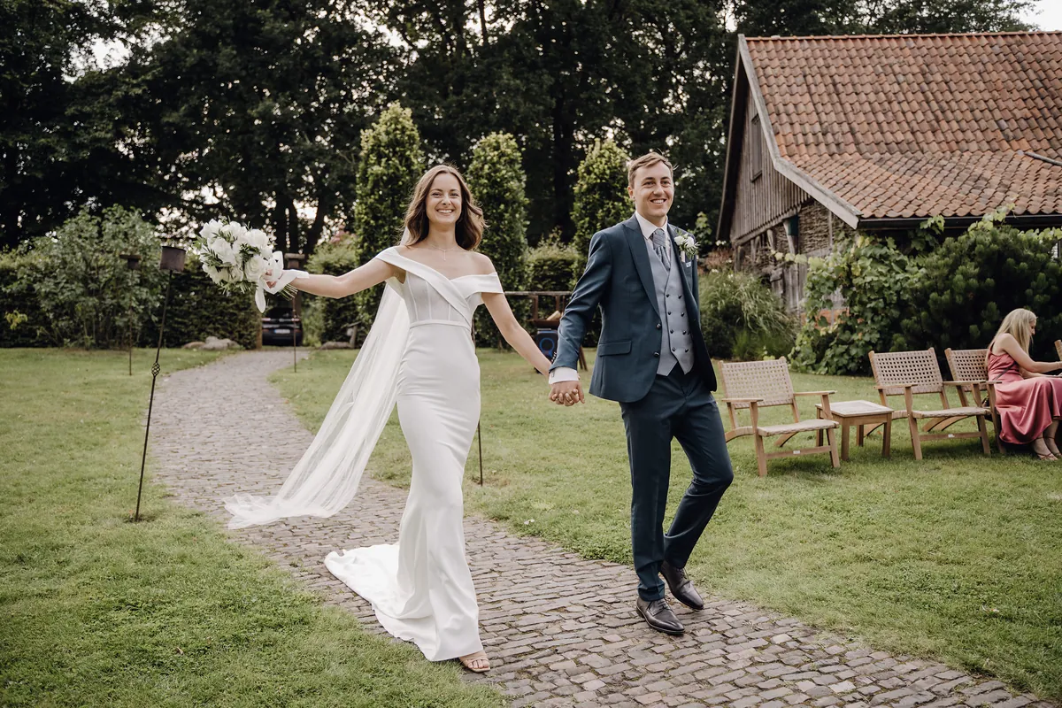 Luisa und Jonas kommen lachend Hand in Hand über einen gepflasterten Gartenweg, sie streckt den Brautstrauß zur Seite, ihr langer Schleier weht, rechts ein altes Holzgebäude mit Ziegeldach.
