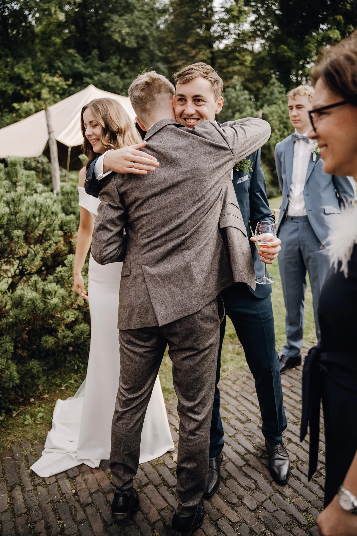 Ein Gast im graubraunen Anzug umarmt Jonas, der ein Glas in der Hand hält und breit über dessen Schulter lächelt, links steht Luisa lächelnd mit gesenktem Blick, rechts weitere Gäste.