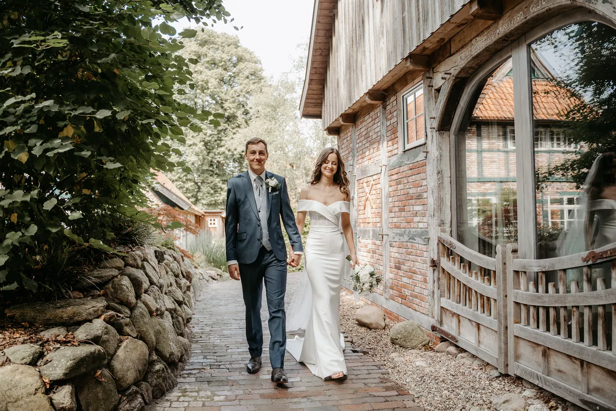 Luisa und Jonas gehen Hand in Hand den Klinkerweg zwischen Feldsteinmauer und Fachwerkhaus entlang, er blickt in die Kamera, sie lächelnd nach unten; im Rundbogenfenster spiegelt sich ein Fachwerkhaus mit rotem Ziegeldach.