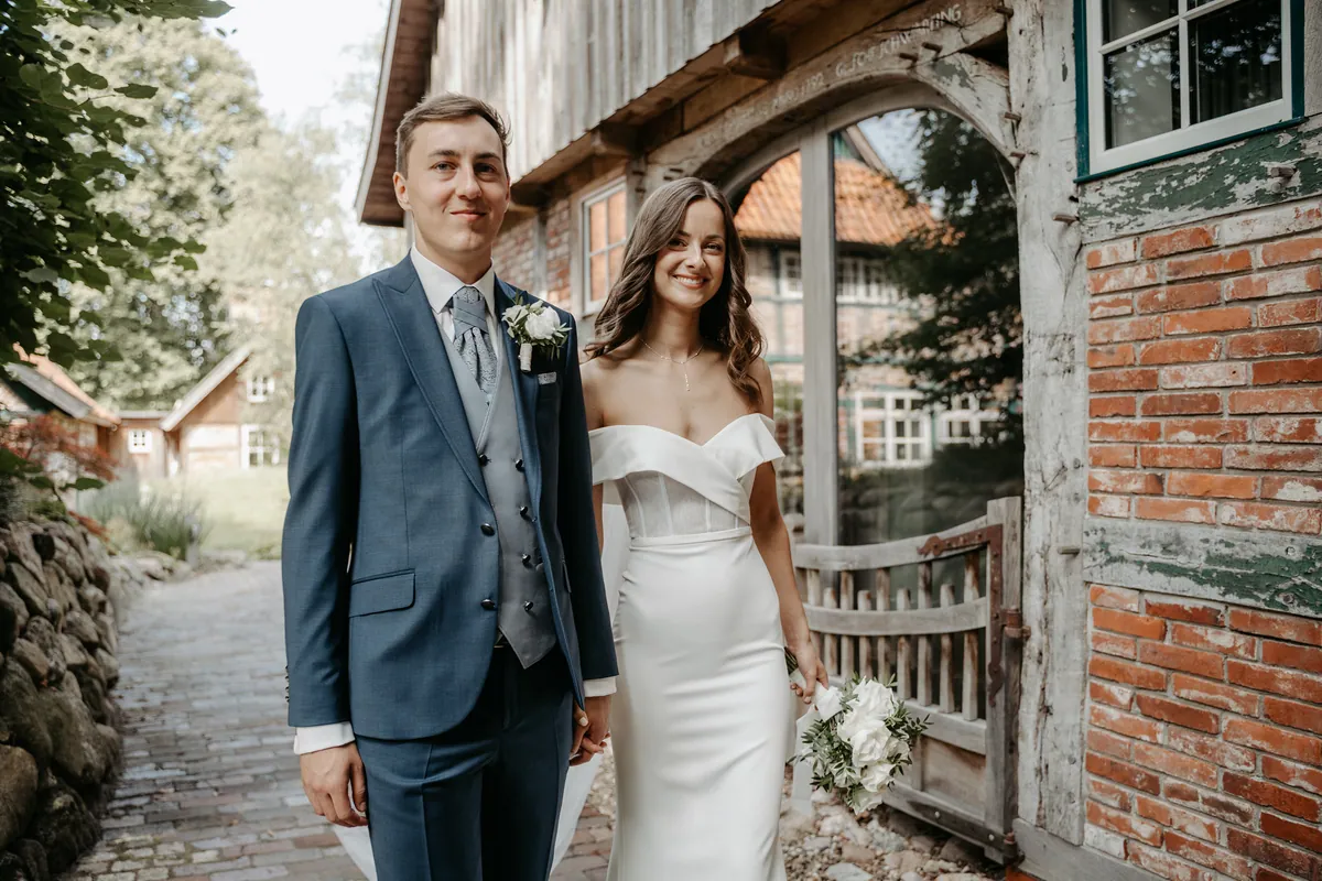 Jonas und Luisa stehen Hand in Hand vor dem verglasten Rundbogen des Fachwerkhauses und schauen in die Kamera, er etwas vor ihr, sie lächelt breit und hält den Rosenstrauß seitlich am Kleid.