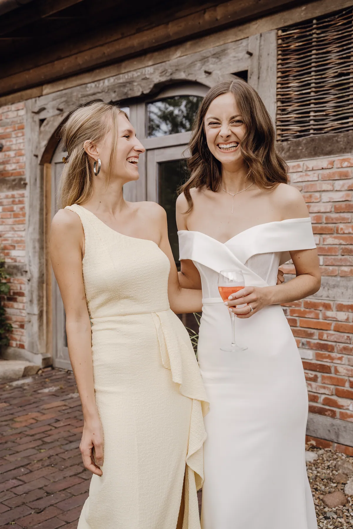 Luisa und eine blonde Frau im blassgelben One-Shoulder-Kleid lachen eng beieinander vor einer Fachwerkwand aus alten Ziegeln, Luisa hält ein Glas mit rosafarbenem Getränk.