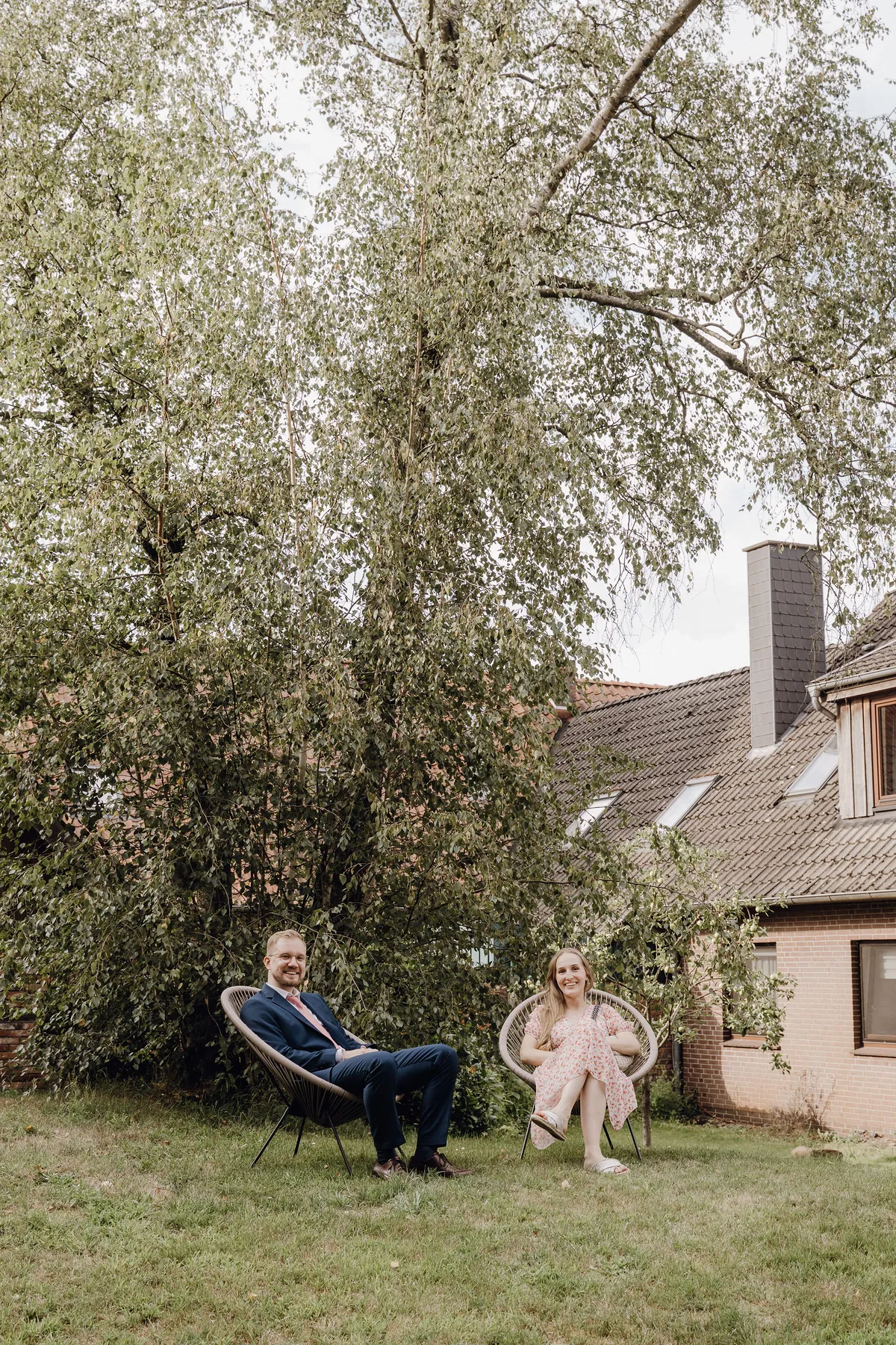 Ein Mann mit Brille im blauen Anzug mit rosa Krawatte und eine Frau im geblümten Kleid sitzen lächelnd in runden Gartensesseln auf dem Rasen unter einer hohen Birke, rechts ein Backsteinhaus.