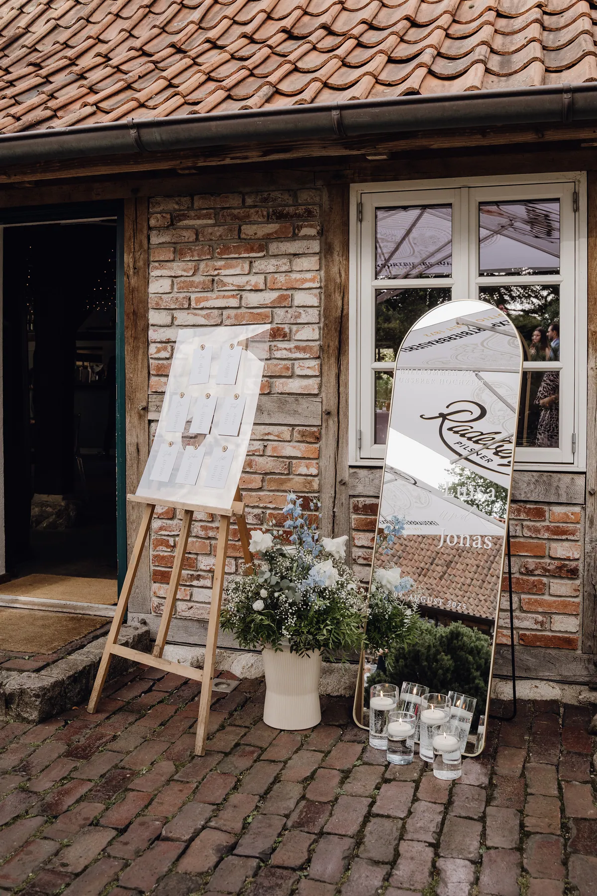 Vor einer Fachwerkwand mit Backsteinen stehen eine Staffelei mit Sitzplan auf Acryl, ein üppiges Gesteck aus weißen Rosen, Schleierkraut und hellblauem Rittersporn sowie ein Bogenspiegel mit den Namen Luisa und Jonas, davor Schwimmkerzen.