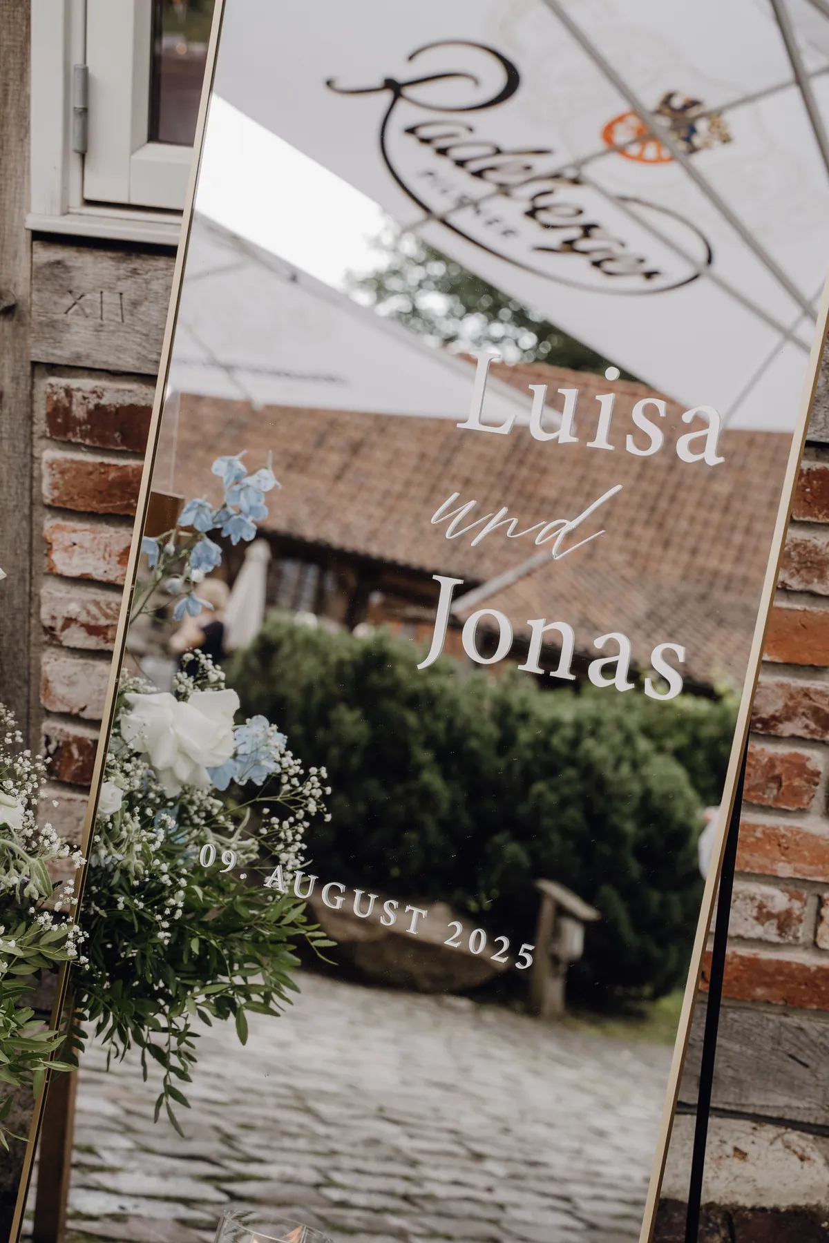 Auf dem goldgerahmten Spiegel stehen in weißer Schrift «Luisa und Jonas» und «09. AUGUST 2025», darin spiegeln sich ein Sonnenschirm, ein Ziegeldach und eine Hecke, links hellblauer Rittersporn und weiße Rosen.