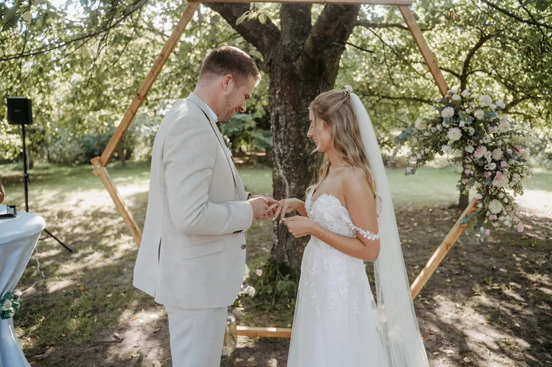Ringtausch am sechseckigen Holzbogen mit Rosen und Eukalyptus: Luca steckt Maira lächelnd den Ehering an.