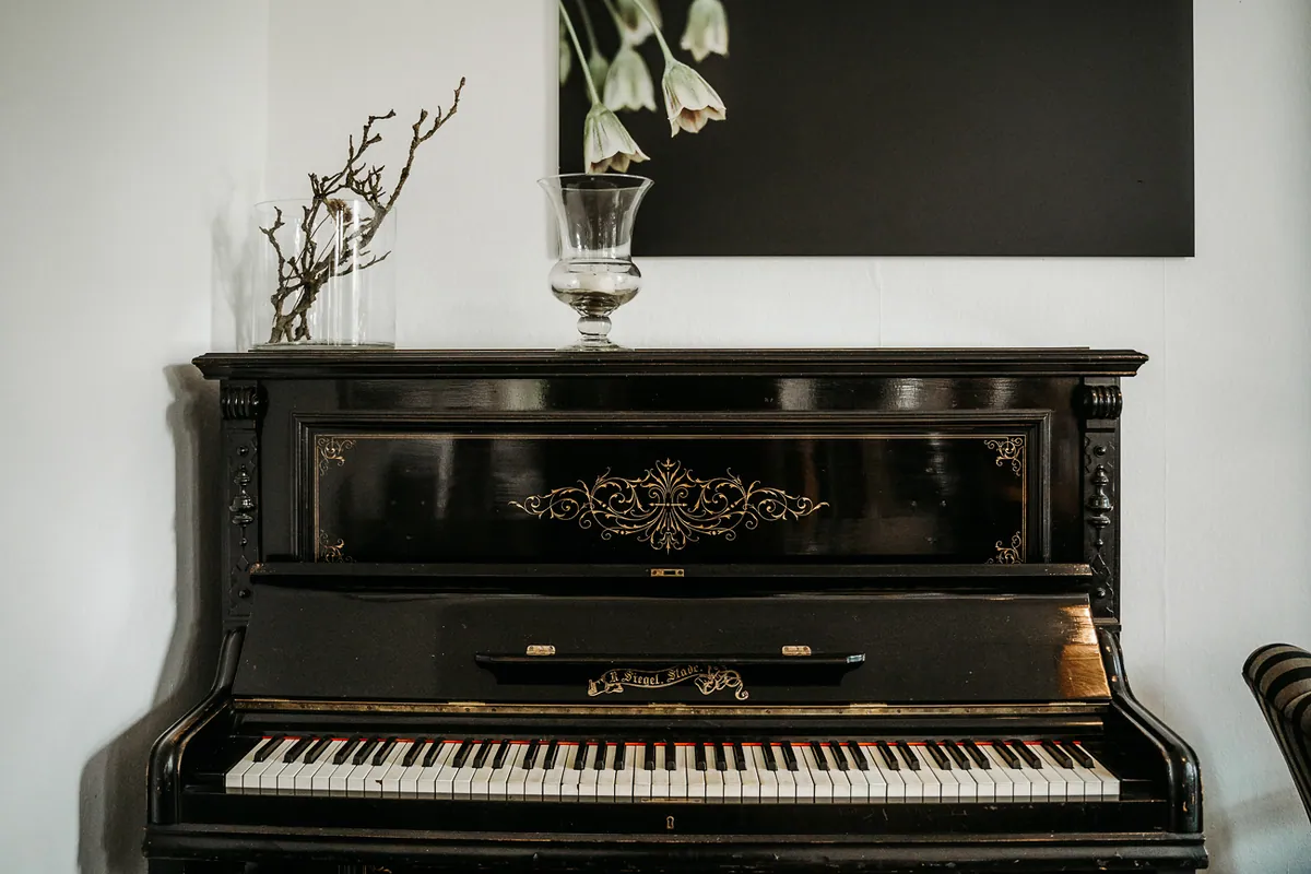 Ein altes schwarzes Klavier mit goldenen Verzierungen steht vor weißer Wand, darauf ein Glaspokal und eine Vase mit kahlen Zweigen.