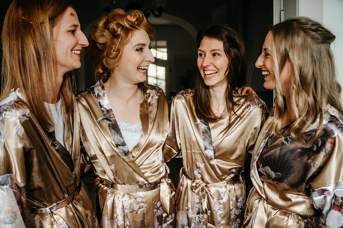 Nadin steht lachend mit drei Frauen eng beieinander, alle vier tragen goldene Satin-Morgenmäntel mit Blumenmuster.