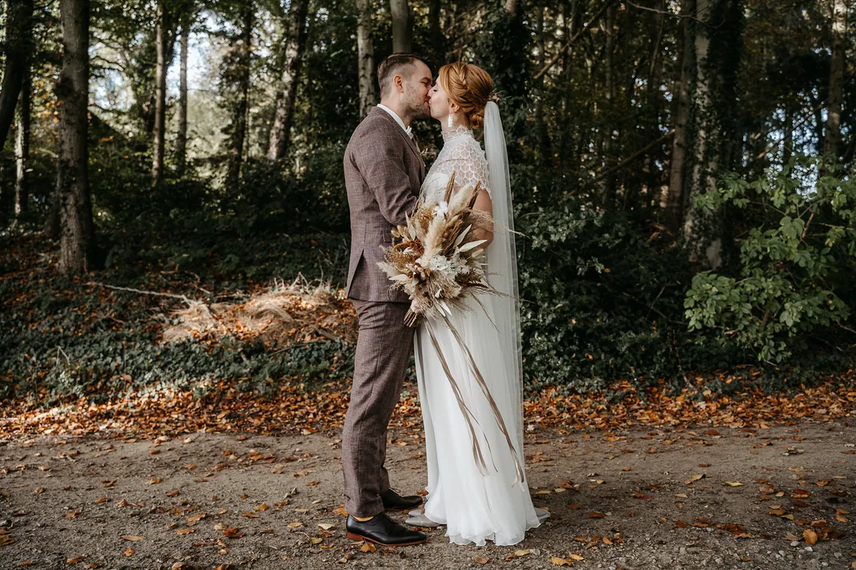 Nadin und Kai küssen sich im herbstlichen Wald, sie hält einen Strauß aus Trockenblumen
