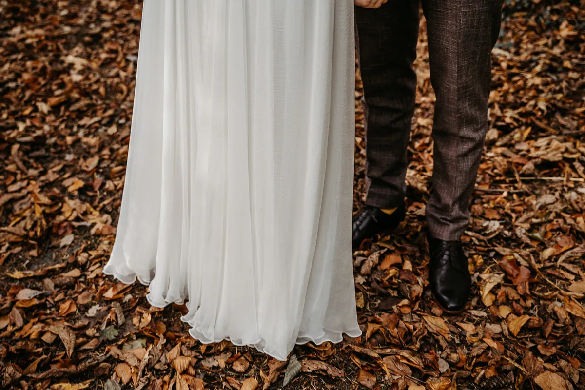 Detail auf Herbstlaub: der fließende Saum des Brautkleids neben den Beinen des Bräutigams in Karohose und schwarzen Lederschuhen.