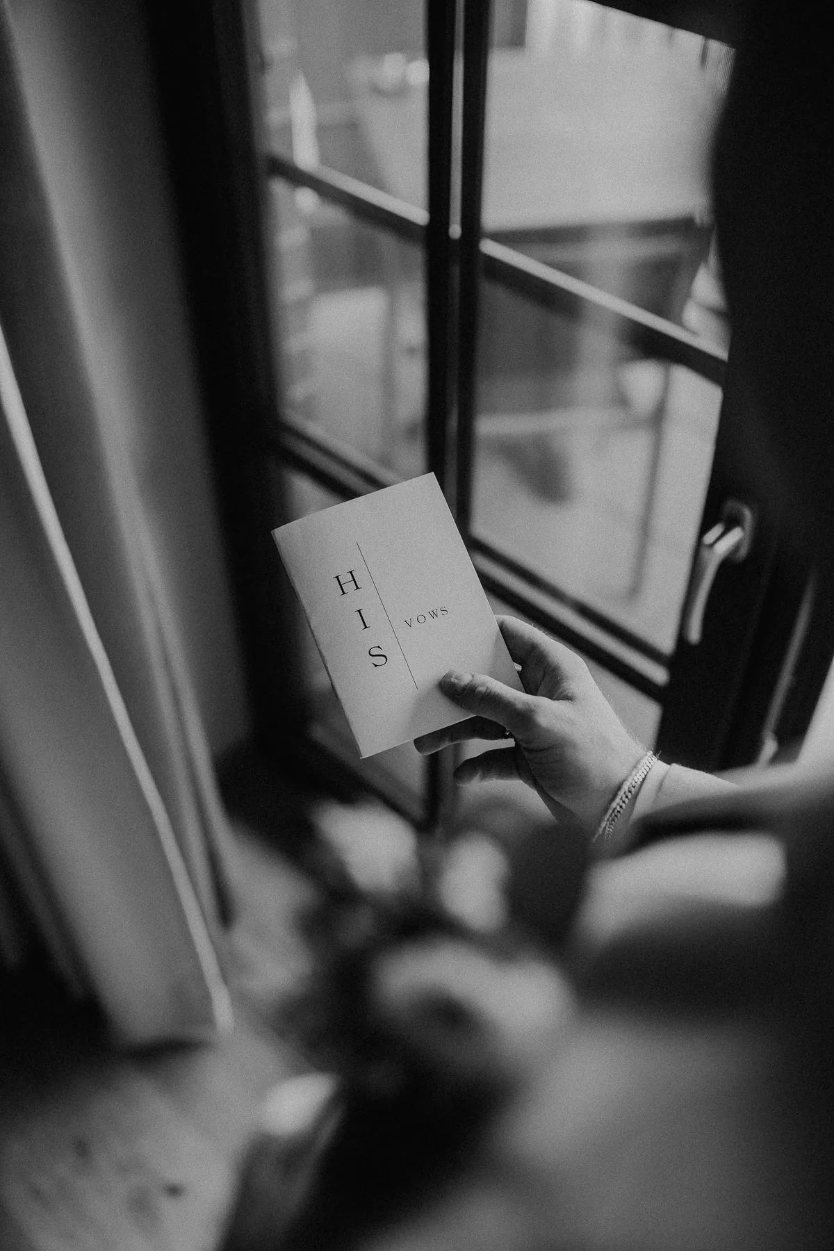 Schwarzweißaufnahme: Leon hält am Fenster ein kleines weißes Heft mit der Aufschrift «HIS VOWS», am Handgelenk ein Gliederarmband, am Finger ein Ring.