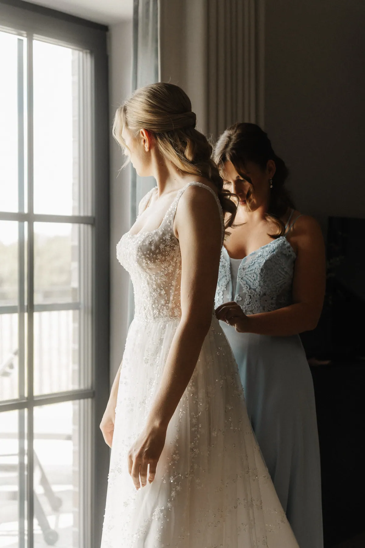 Neele steht im glitzernden Brautkleid am Fenster und blickt hinaus, hinter ihr schließt eine Frau mit dunklem Haar im hellblauen Spitzenkleid das Kleid am Rücken.