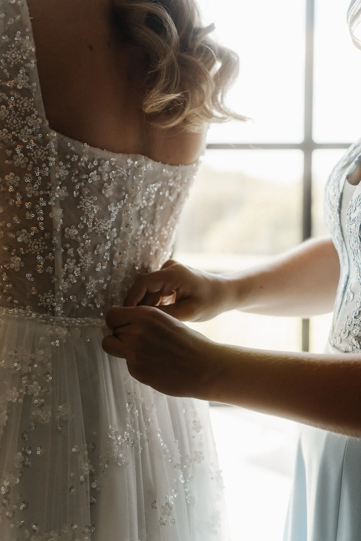 Nahaufnahme am Fenster: Zwei Hände schließen die Knöpfe am Rücken der paillettenbestickten Corsage, darüber fallen blonde Locken, rechts der Spitzenstoff eines hellblauen Kleides.