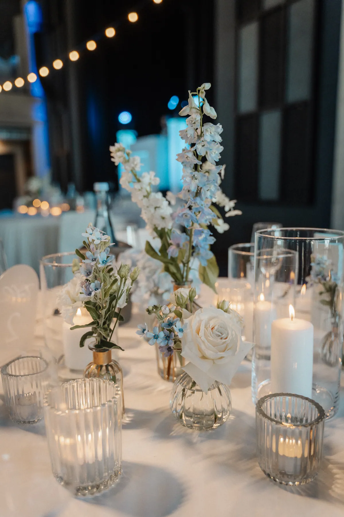 Auf dem weiß gedeckten Tisch stehen kleine Glasvasen mit weißer Rose, Rittersporn und hellblauen Sternblüten zwischen Stumpenkerzen und geriffelten Windlichtern, darüber verschwimmt eine Lichterkette.