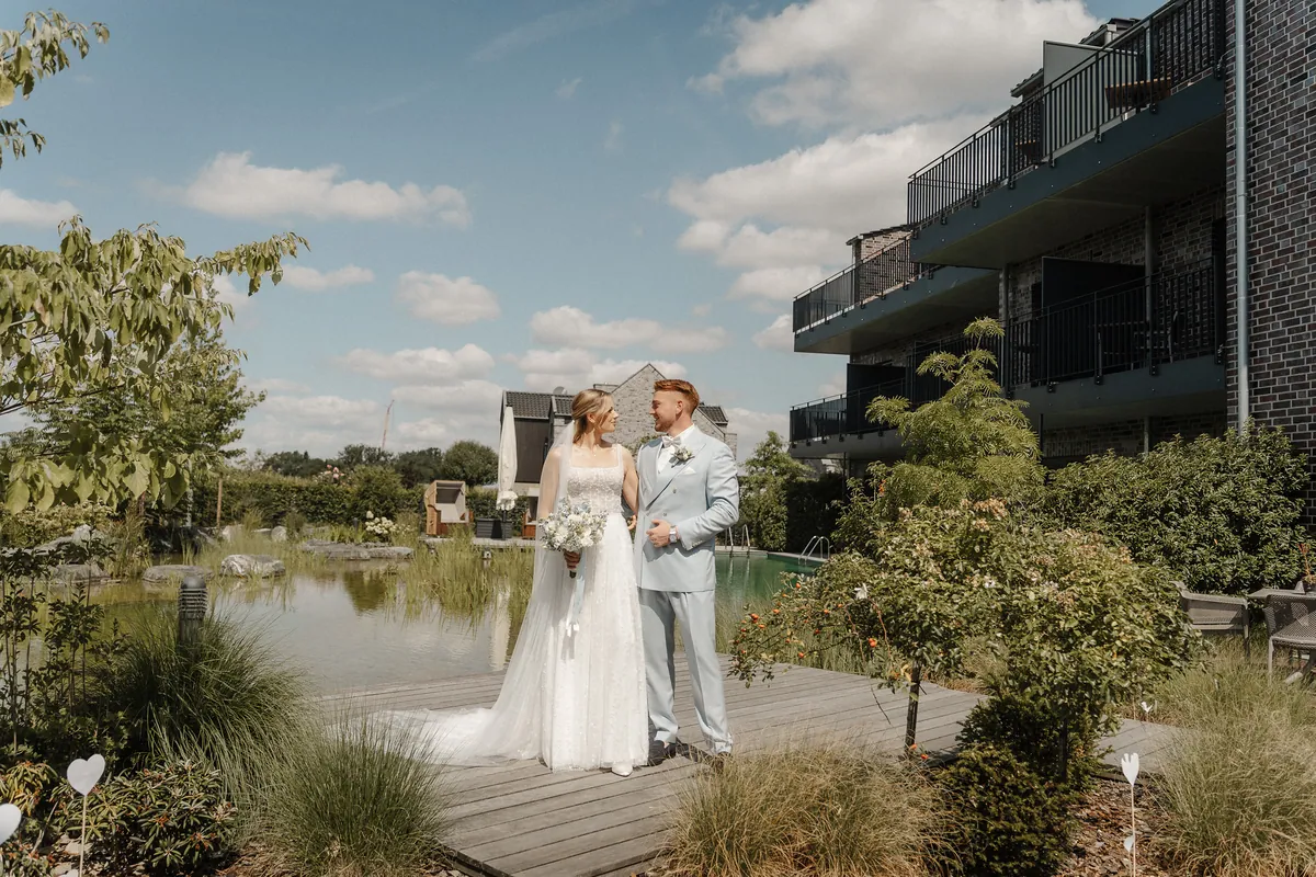 Neele und Leon stehen auf dem Holzsteg zwischen Naturteich und Klinkerbau mit Balkonen und schauen sich an, sie hält den hellblau-weißen Strauß, vorn Gräser und weiße Herzstecker.