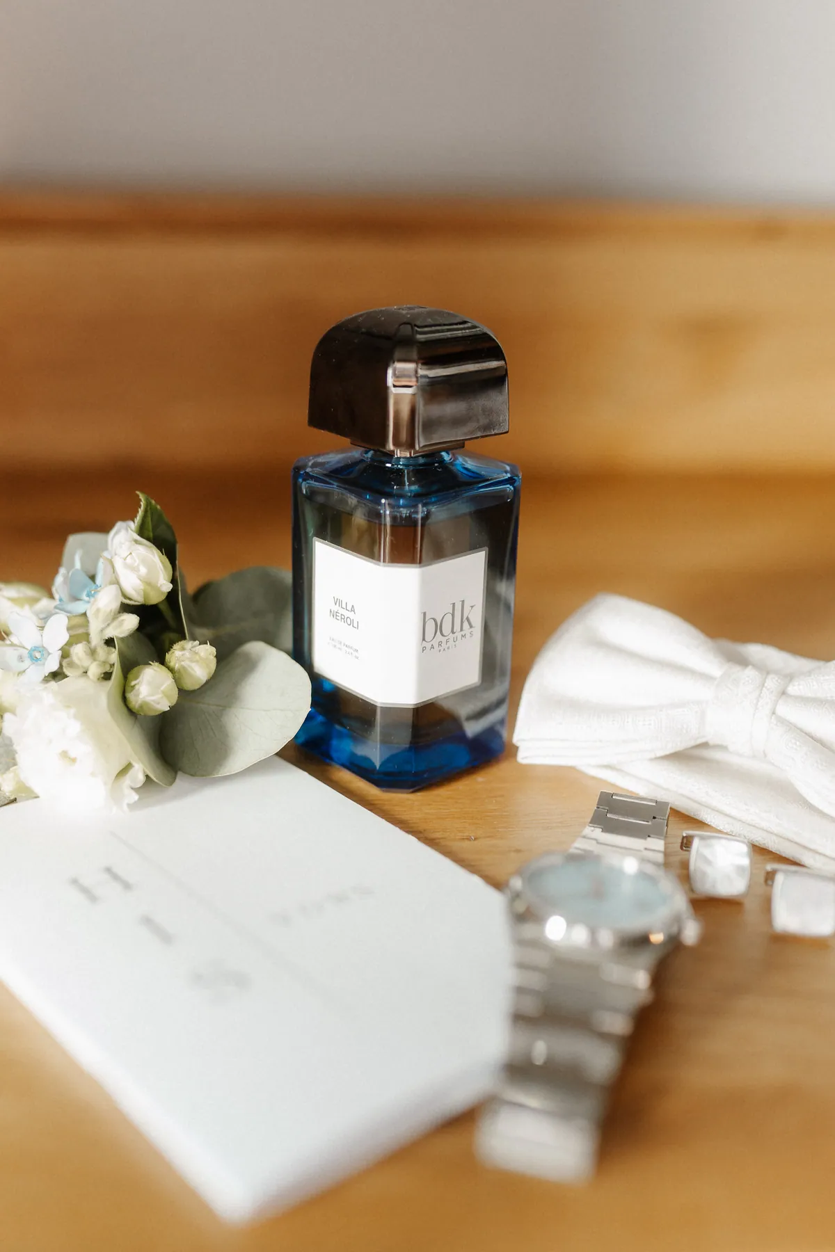 Auf einem Holztisch liegen ein blauer Parfumflakon, eine weiße Fliege mit Jacquardmuster, eine Stahluhr mit hellblauem Zifferblatt, Manschettenknöpfe mit Perlmutt und ein Anstecksträußchen aus weißen Röschen, hellblauen Blüten und Eukalyptus.