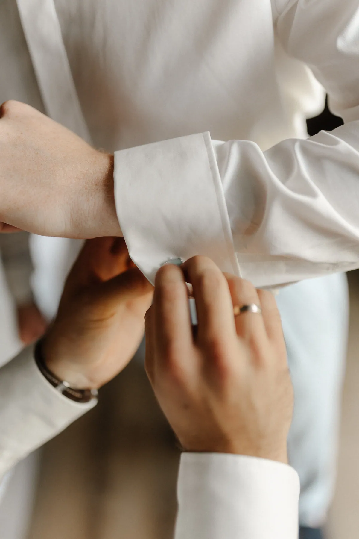 Zwei Hände, eine mit Ring, die andere mit Lederarmband, schließen einen hellblau schimmernden Manschettenknopf an der weißen Hemdmanschette eines sommersprossigen Handgelenks.