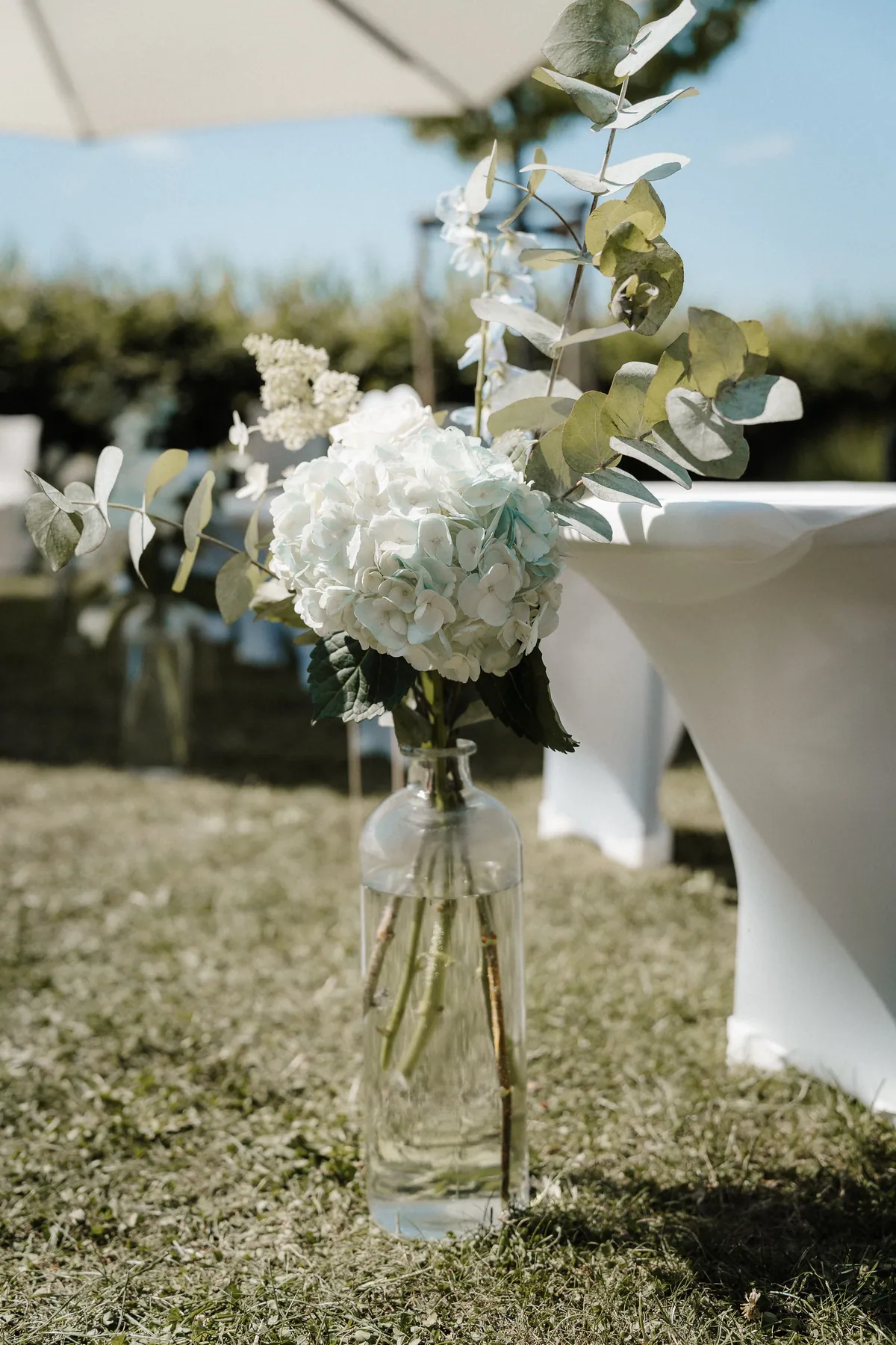 Eine weiß-hellblaue Hortensie mit Eukalyptuszweigen und weißen Blütenrispen steht in einer Glasflasche im Gras neben einer weiß bezogenen Bank, oben der Saum des Sonnenschirms.