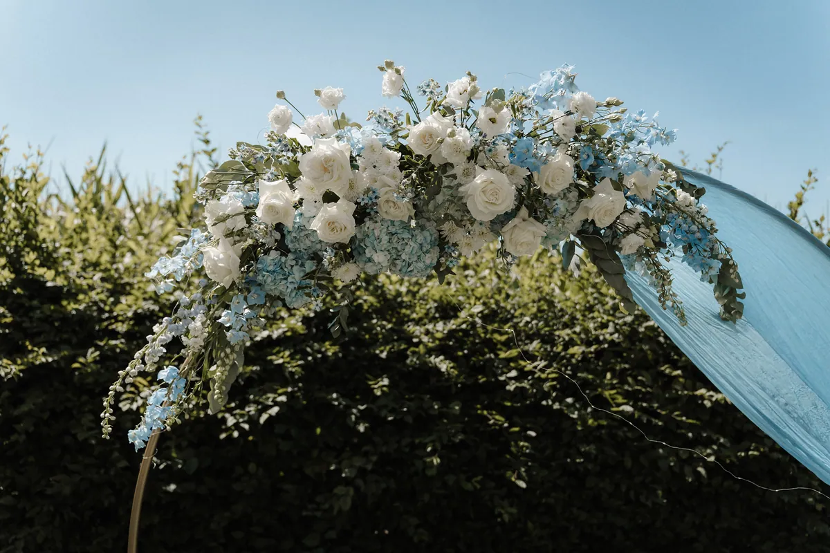 Vor blauem Himmel und dunkler Hecke trägt der goldene Traubogen ein üppiges Gesteck aus weißen Rosen, hellblauen Hortensien und Rittersporn, rechts fällt ein hellblaues Tuch herab.