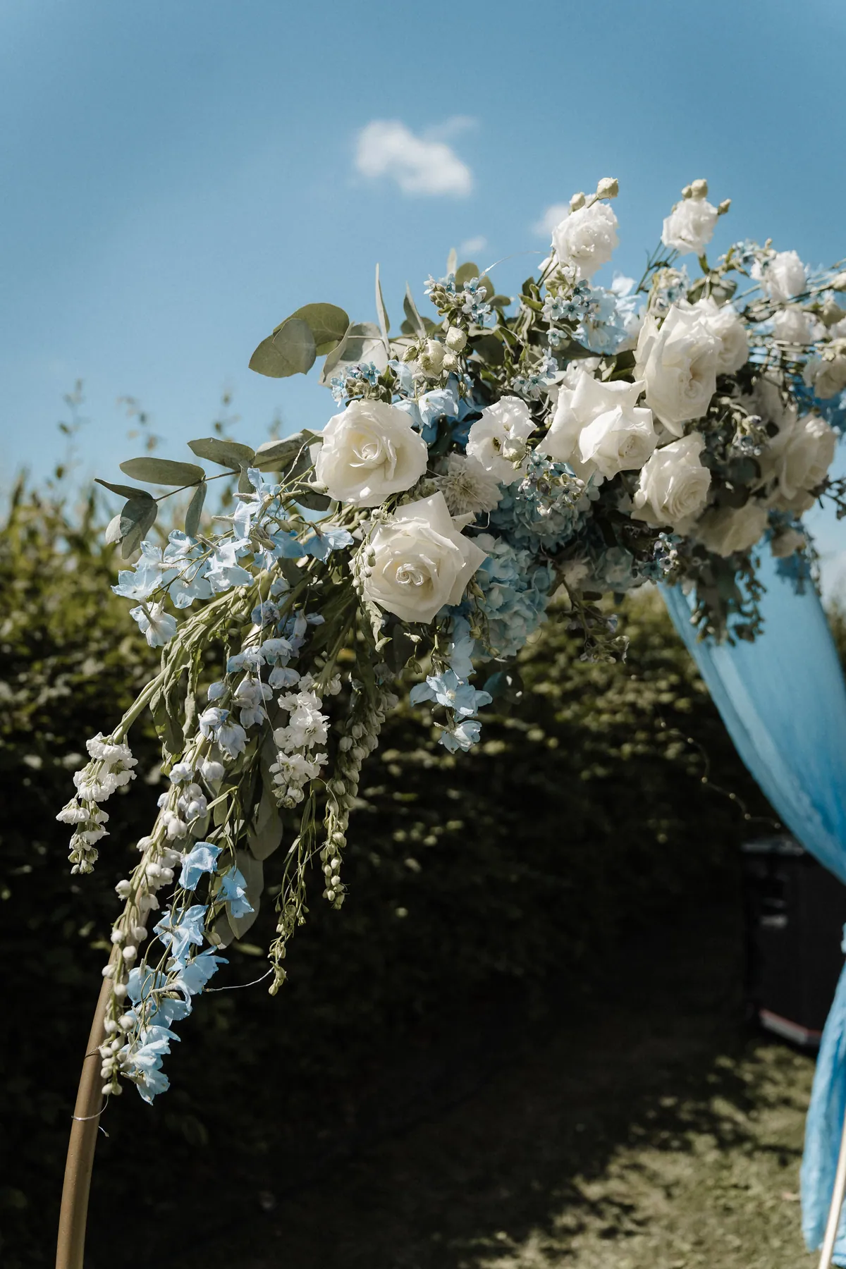 Nahaufnahme der linken Seite des Traubogens: Weiße Rosen, Eustoma, hellblauer Rittersporn und Eukalyptus ranken am goldenen Rundbogen herab, darüber blauer Himmel mit einer kleinen Wolke.