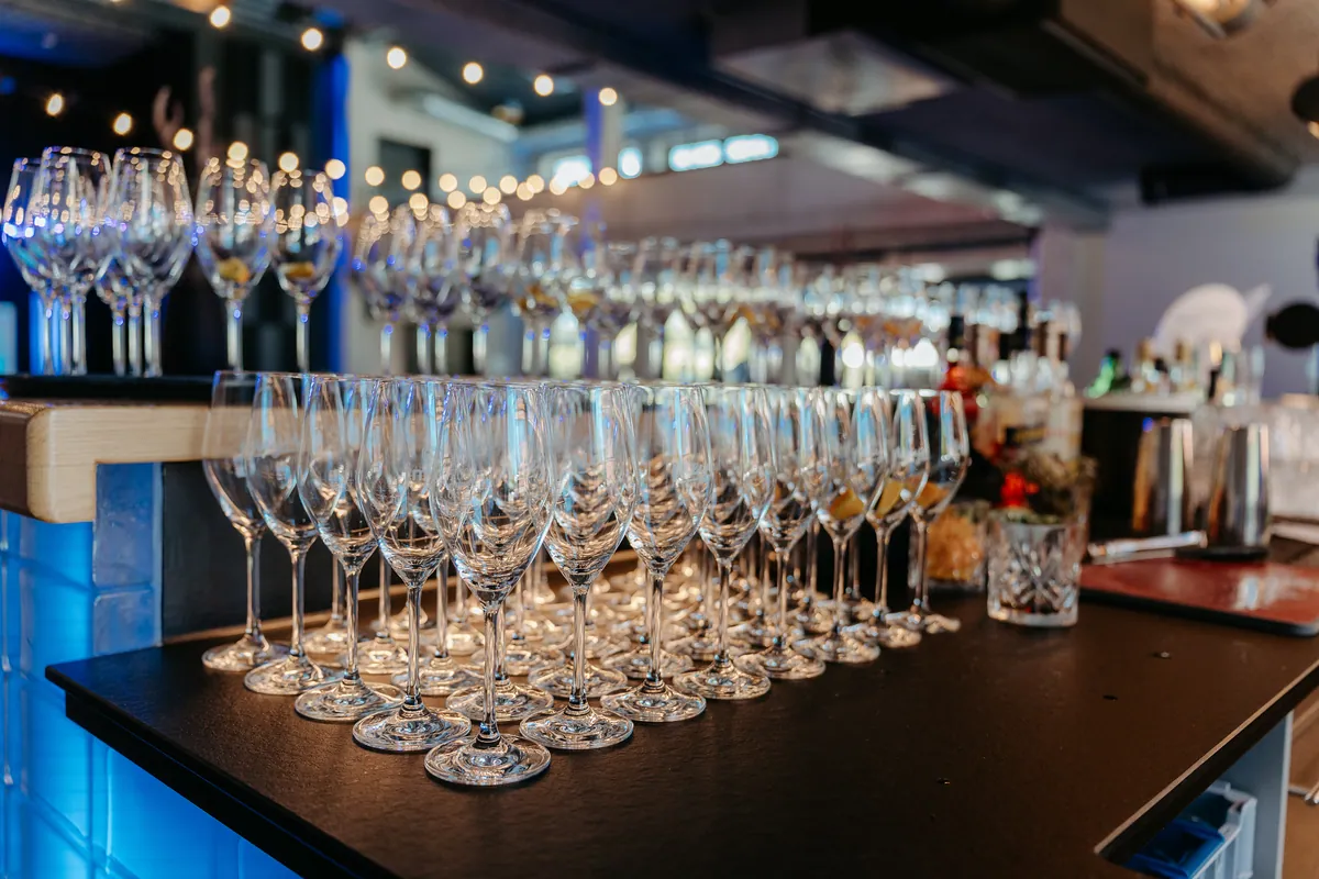 Auf einer schwarzen Theke stehen dicht an dicht leere Sekt- und Weingläser, einige mit Zitrusscheibe, dahinter Flaschen, Lichterketten und blau leuchtendes Licht an der Bar.