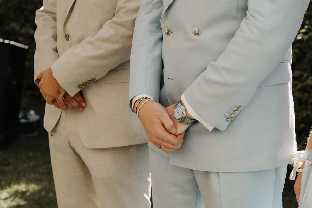 Nahaufnahme der Hände: Leon umfasst im hellblauen Zweireiher sein Handgelenk mit Uhr, an der anderen Hand Ring, Ketten- und Perlenarmband, links die gefalteten Hände des Mannes im beigen Anzug.