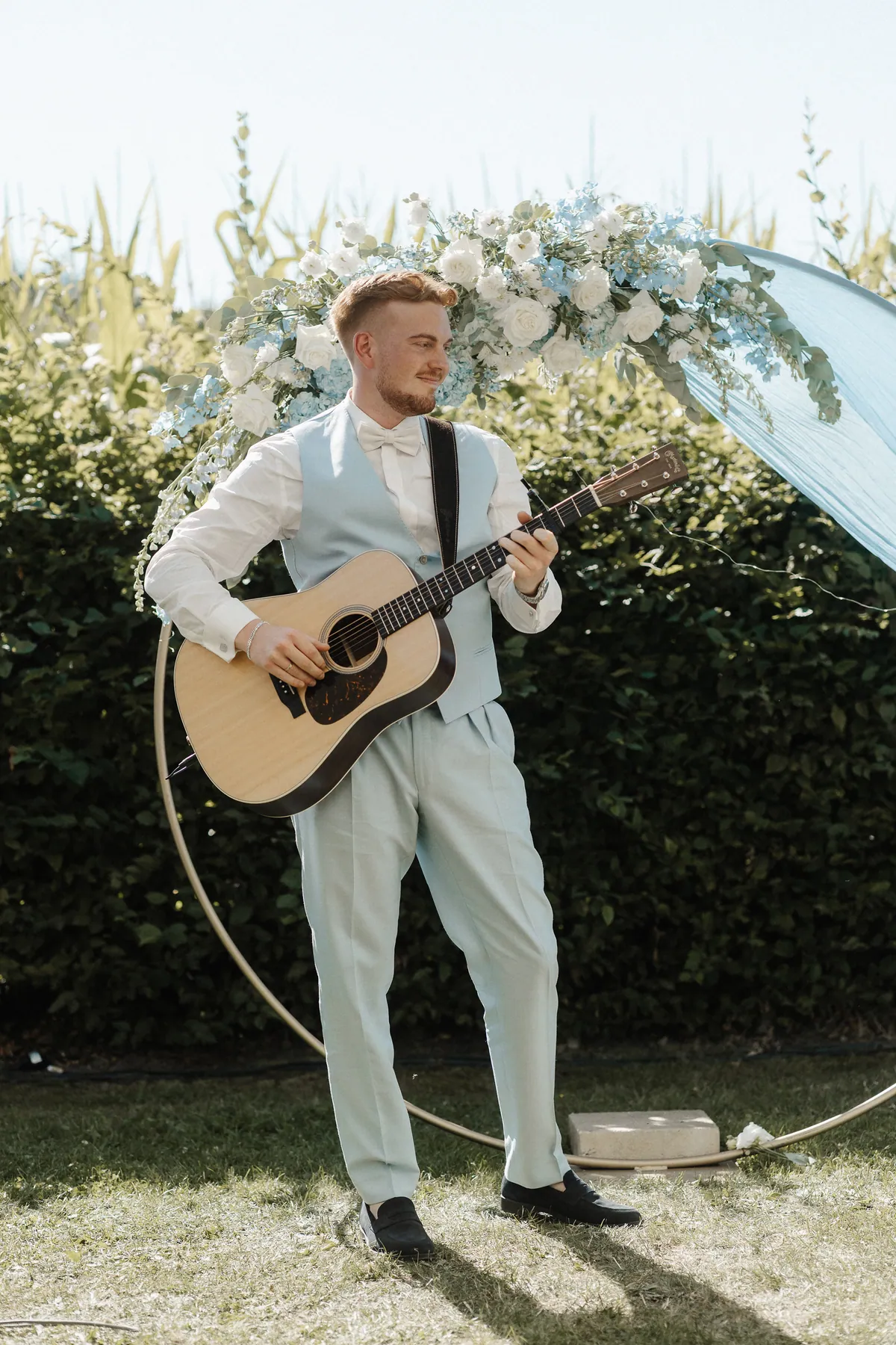 Leon steht ohne Sakko in hellblauer Weste und weißer Fliege vor dem Blumenring aus weißen Rosen und hellblauem Rittersporn und spielt lächelnd auf einer Akustikgitarre.