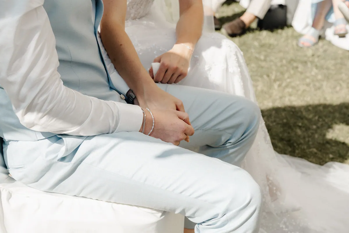 Nahaufnahme im Sitzen: Neele hat sich bei Leon eingehakt, ihre Hand liegt auf seiner, an seinem sommersprossigen Handgelenk Silberkette und Perlenarmband, in der anderen Hand hält sie ein Taschentuch über dem glitzernden Kleid.