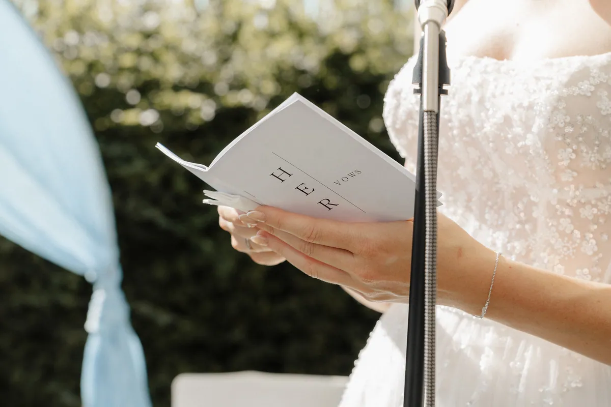 Neele hält ihr Gelübdeheft mit der Aufschrift «HER VOWS» vor dem Mikrofon, die Fingernägel in French-Optik, am Handgelenk ein feines Silberkettchen, links unscharf hellblauer Stoff.