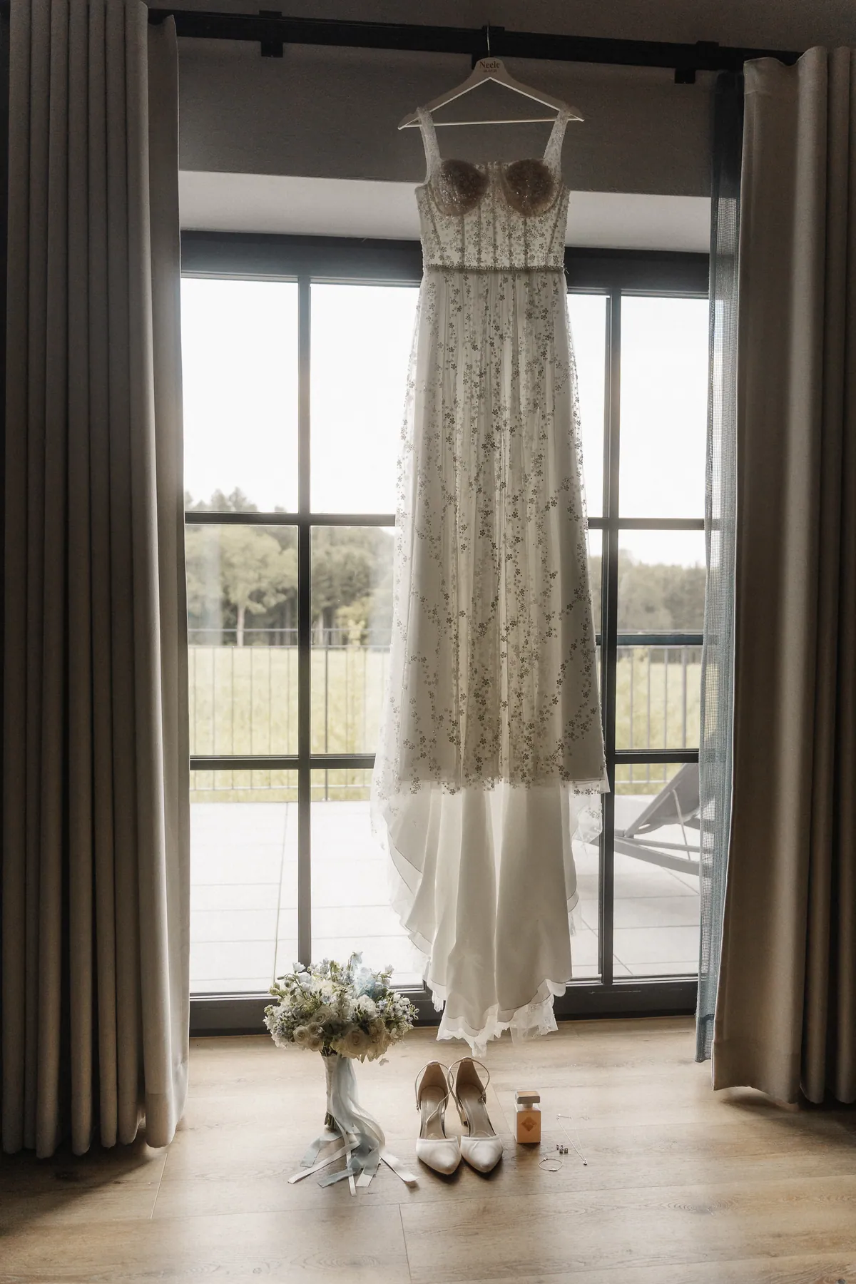 Das Brautkleid mit Pailletten-Blüten hängt auf einem Bügel mit der Aufschrift «Neele 08.08.26» vor einer hohen Fenstertür, davor Brautstrauß, cremefarbene Pumps und ein Parfumflakon.