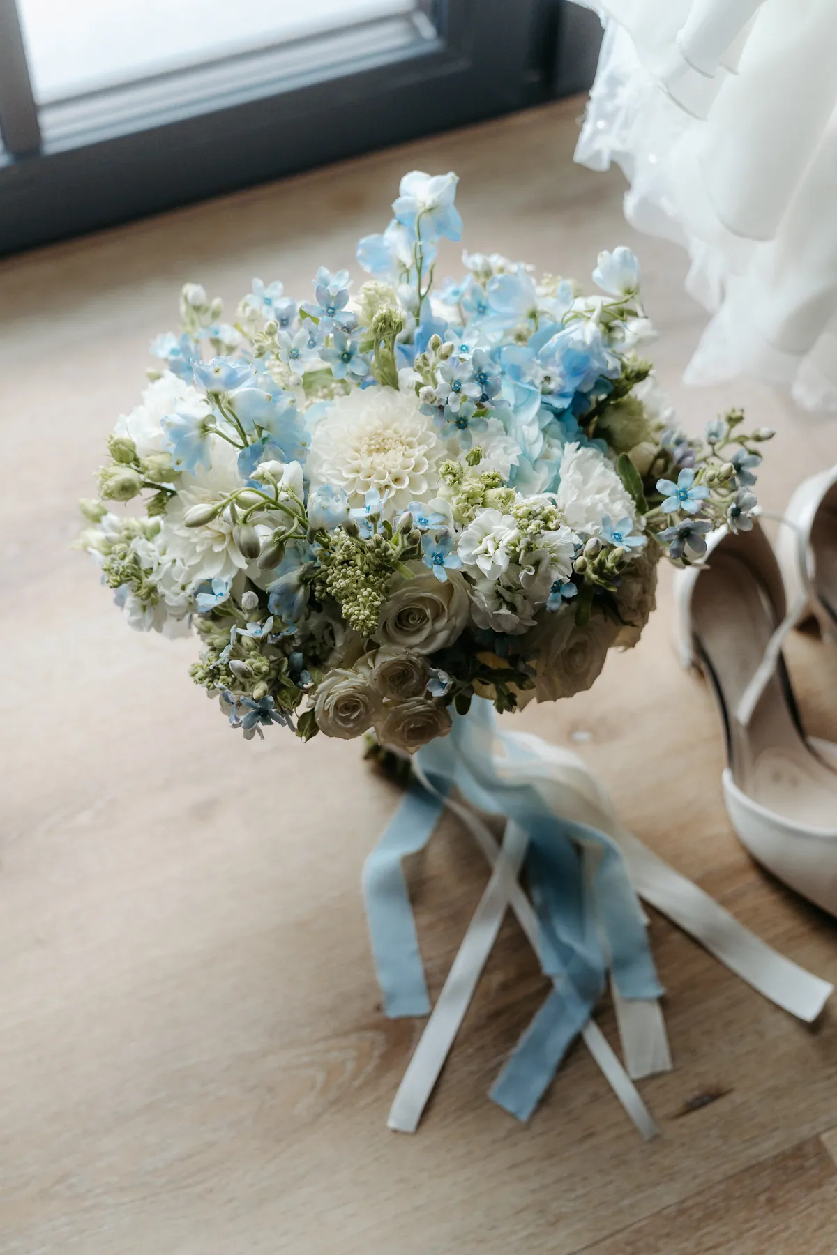 Von oben gesehen steht der Brautstrauß aus weißen Dahlien, Rosen und hellblauen Blüten mit hellblauen und cremefarbenen Bändern auf dem Holzboden, daneben die Brautschuhe und der Saum des Kleides.