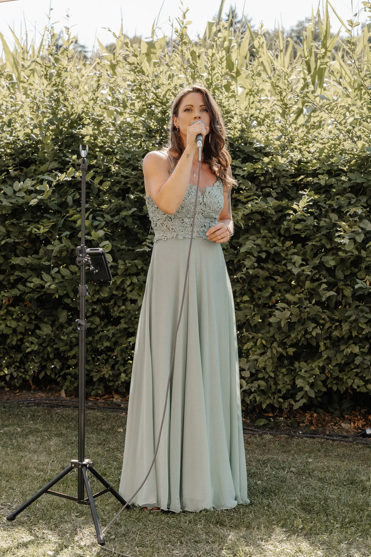 Eine Sängerin im salbeigrünen Kleid mit Spitzenoberteil singt in ein Handmikrofon, neben ihr ein Stativ mit Tablethalterung, hinter ihr Hecke und Maisfeld.