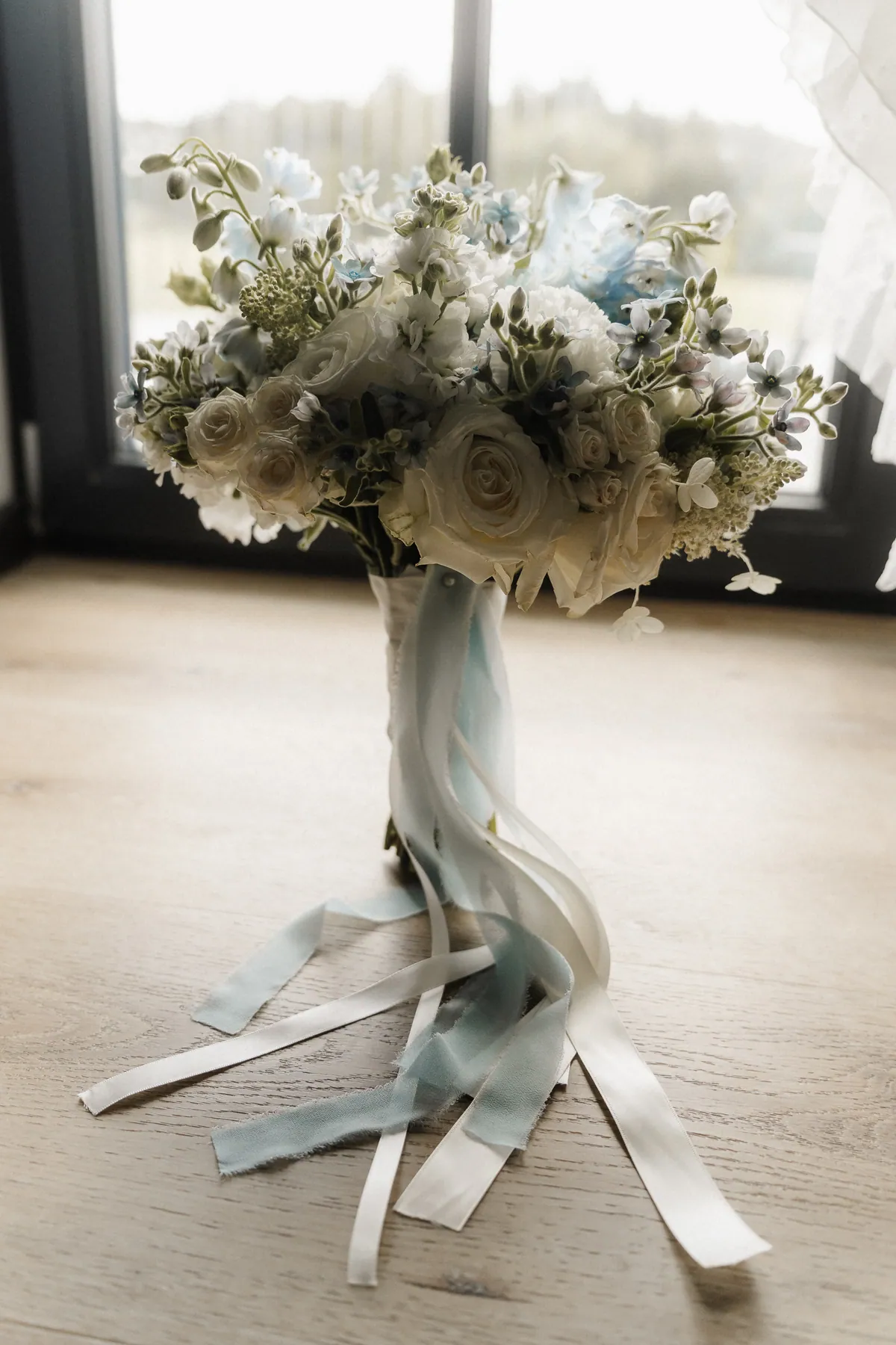 Im Gegenlicht vor der Fenstertür steht der Brautstrauß aus weißen Rosen und hellblauen Blüten aufrecht auf dem Holzboden, die langen Seidenbänder fallen in Schlaufen davor.