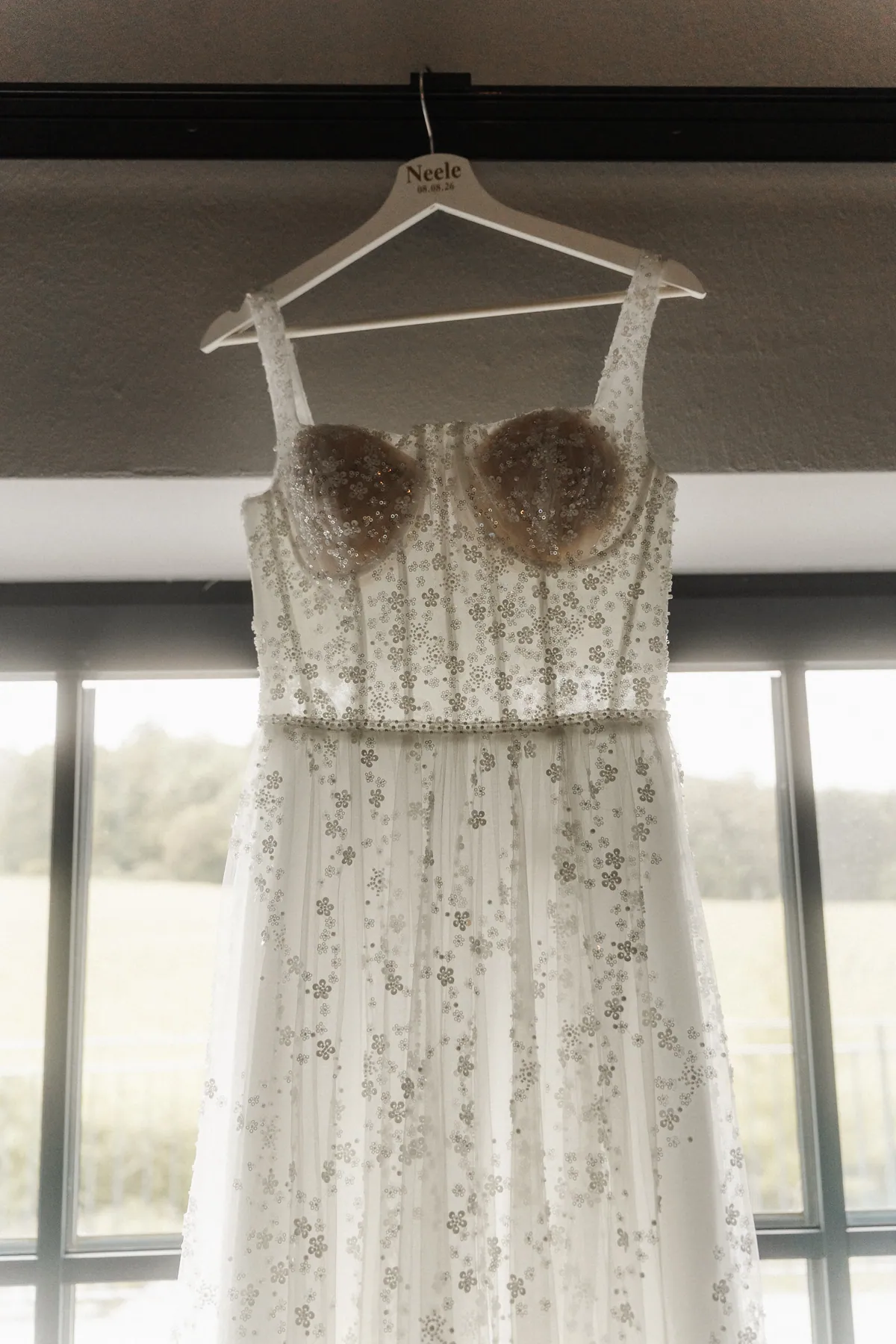 Von unten gesehen hängt das Brautkleid mit Corsage und Pailletten-Blüten auf einem weißen Bügel mit der Aufschrift «Neele 08.08.26» vor dem Fenster mit Blick ins Grüne.