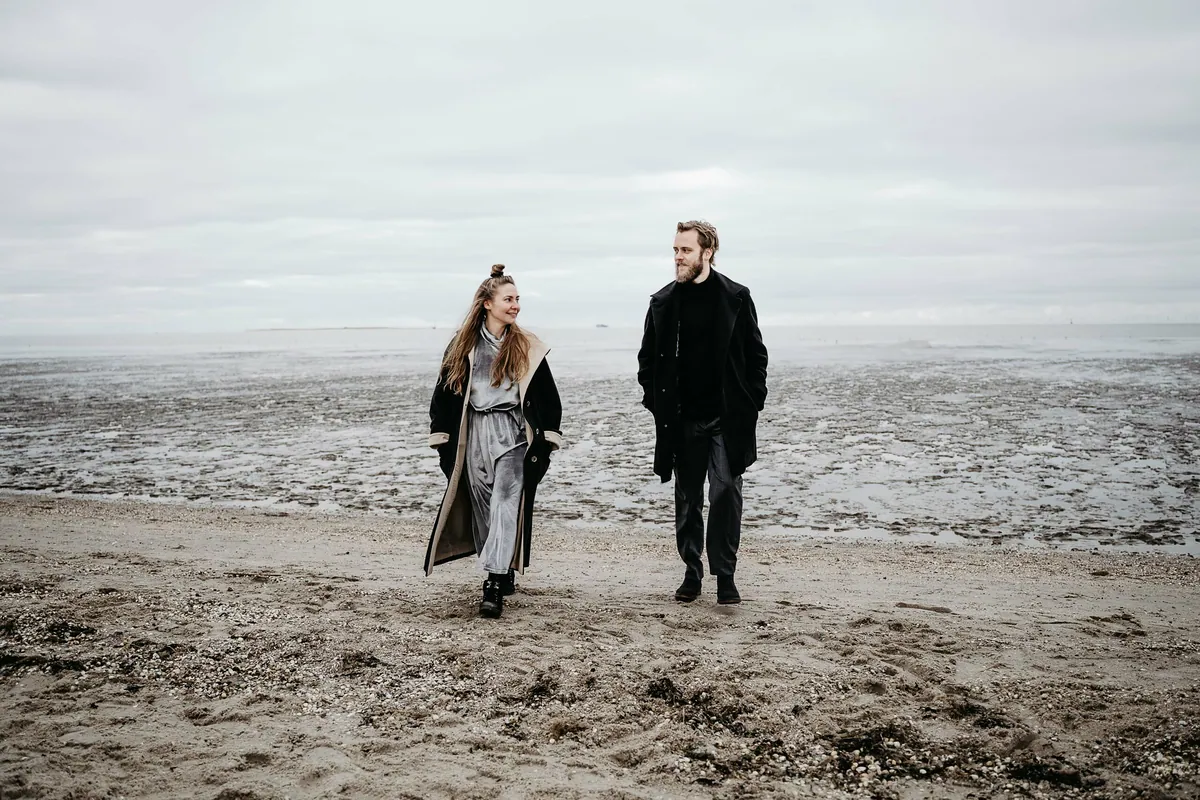 Nebeneinander laufen sie in dunklen Mänteln über den Strand und schauen sich an, hinter ihnen das flache Watt unter tiefen Wolken.
