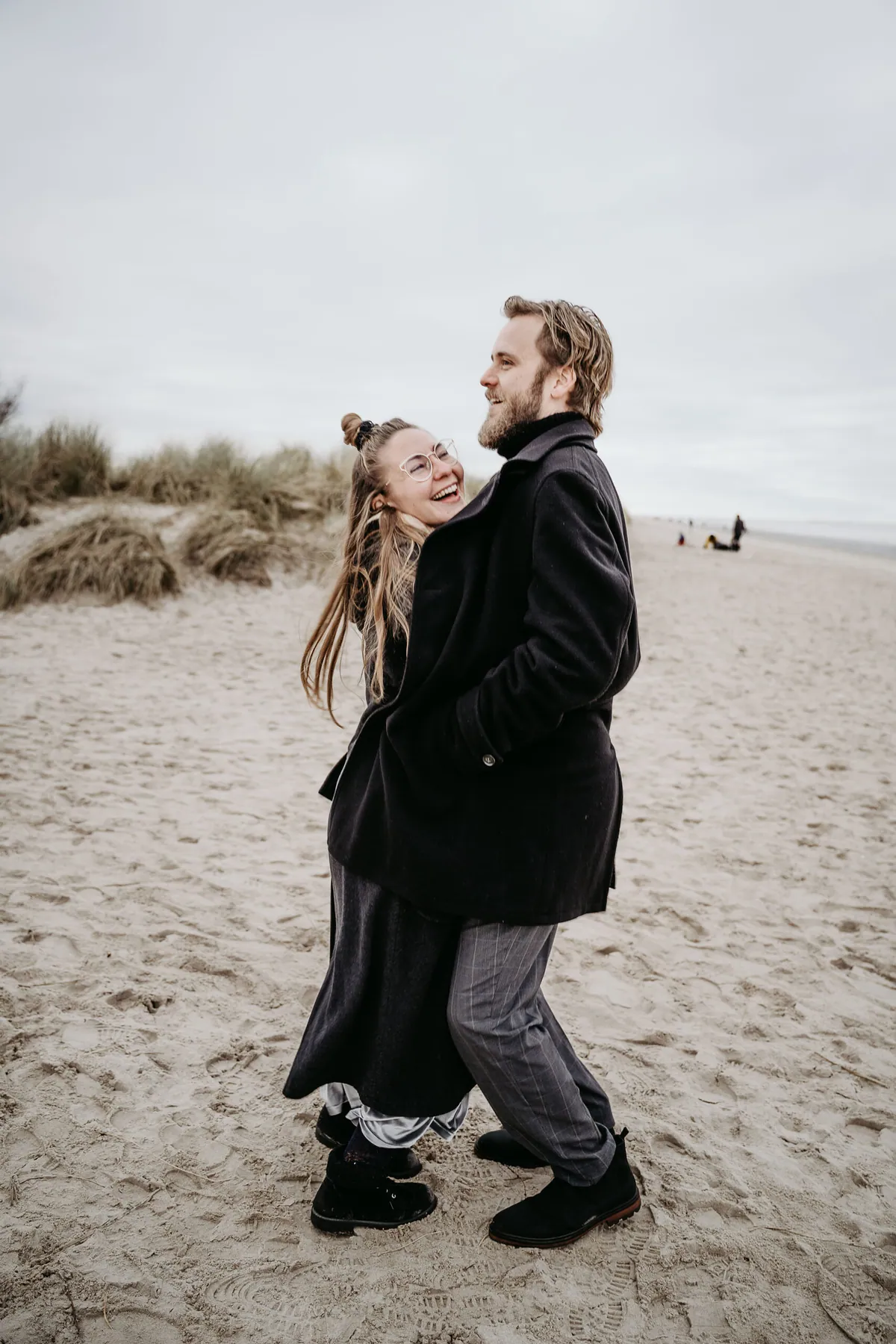 Auf dem Sandstrand vor den Dünen zieht er sie eng an sich, sie lacht mit Brille über seine Schulter; in der Ferne ein paar Spaziergänger.