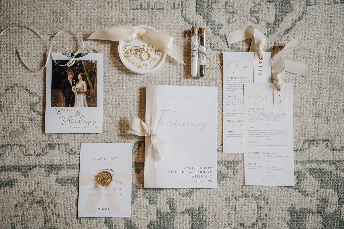 Von oben auf einem Teppich: Einladung mit Paarfoto, Save-the-Date mit Wachssiegel, Ringschale, Menükarten und das Heft zur kirchlichen Trauung mit der Aufschrift «Trauscheune Farminn Wittingen 20.06.2026».
