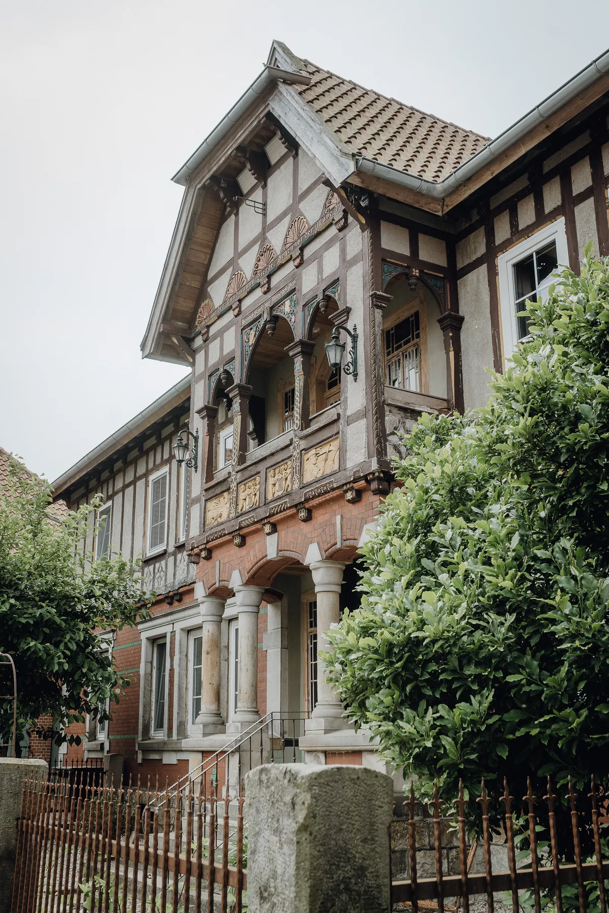 Ein historisches Fachwerkhaus mit geschnitztem Erker, Reliefs und Säulenportal, davor ein rostiges Zaungitter und grüne Büsche unter grauem Himmel.