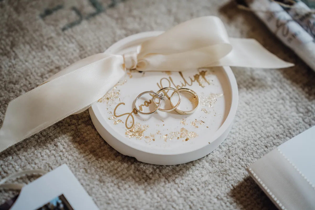 Drei Ringe, einer davon mit kleinem Stein, liegen in einer weißen Schale mit Goldflocken und dem Schriftzug «Sara & Philipp», daneben eine Satinschleife.