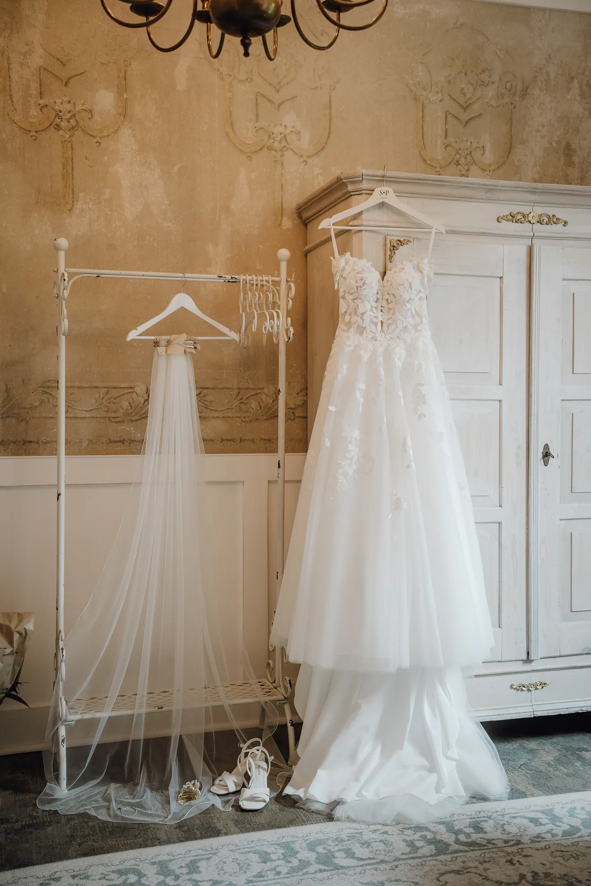 Das Brautkleid mit Blätterspitze hängt am weißen Bauernschrank, daneben ein langer Schleier an einem Kleiderständer, davor stehen weiße Brautschuhe.