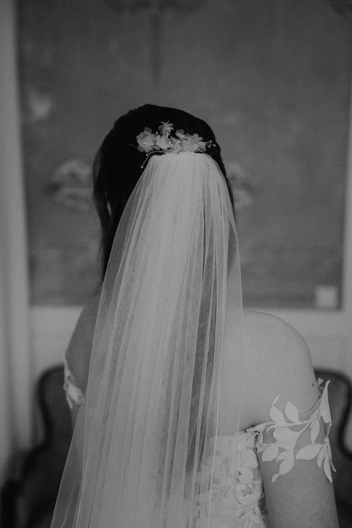 Schwarzweißaufnahme von hinten: Ein Blütenkamm hält den Schleier im Haar der Braut, auf ihrer Schulter liegen Blätter aus Spitze.