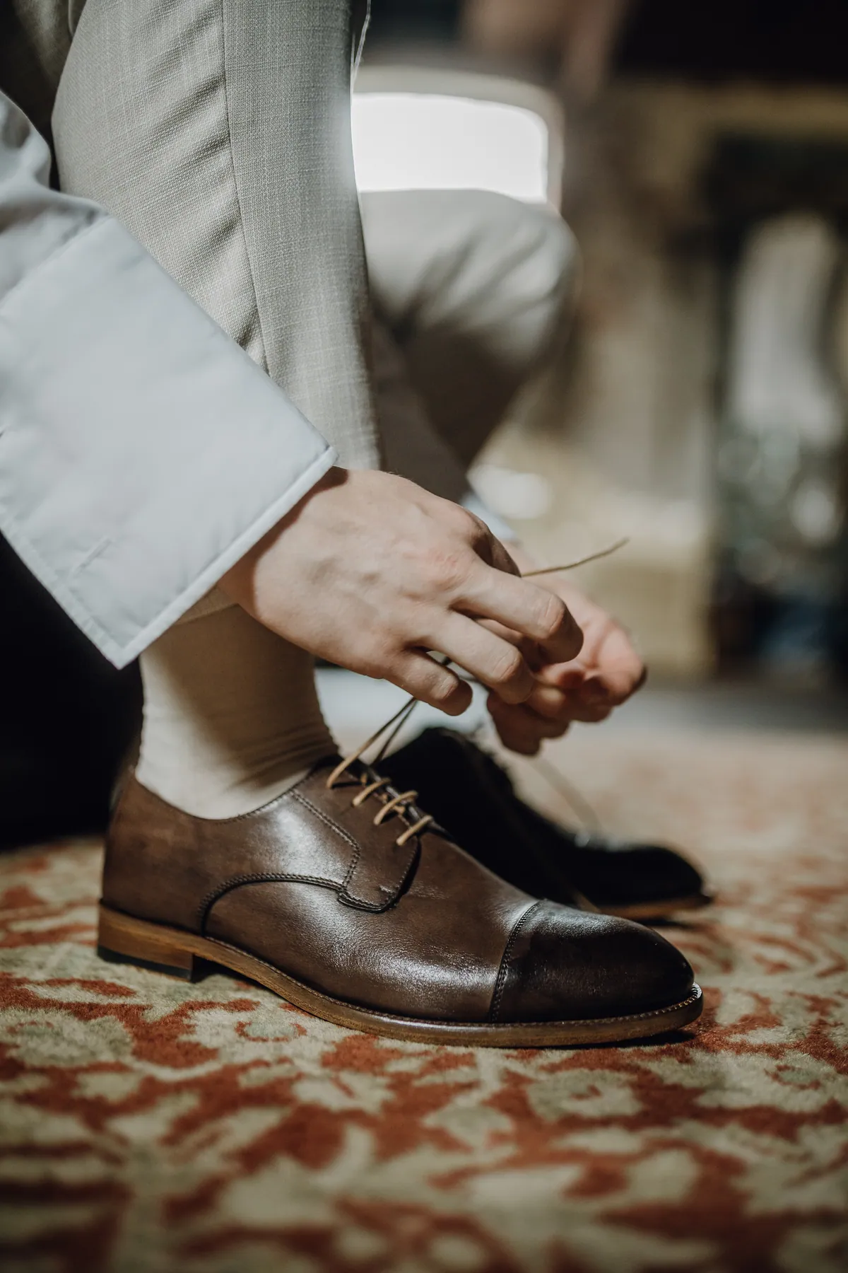 Nahaufnahme: Der Bräutigam schnürt seinen braunen Lederschuh mit dunkler Kappe, dazu helle Hose und beige Socken auf einem rot gemusterten Teppich.